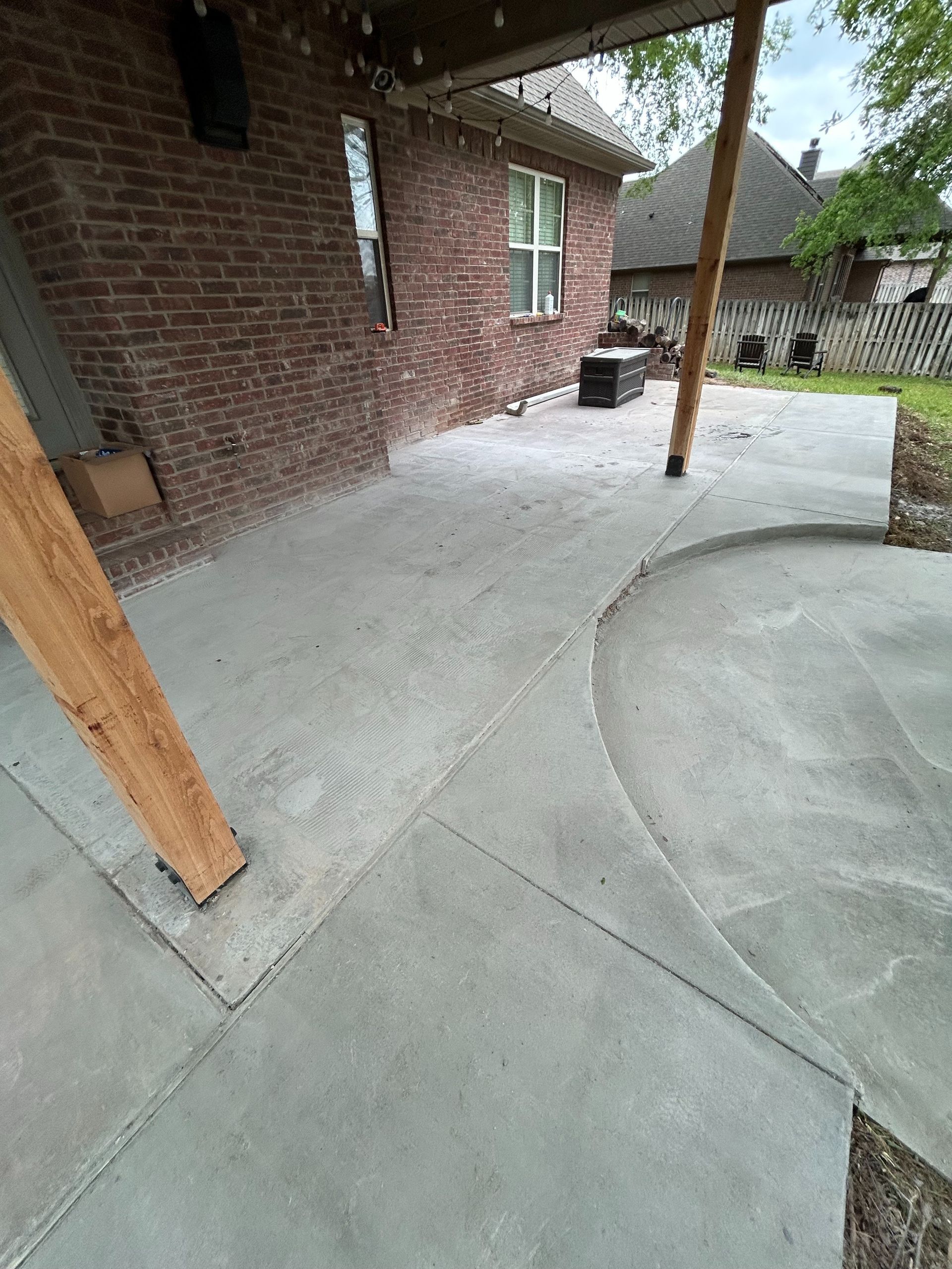 A brick house with a covered concrete patio featuring wooden support posts and a curved edge leading to the yard.