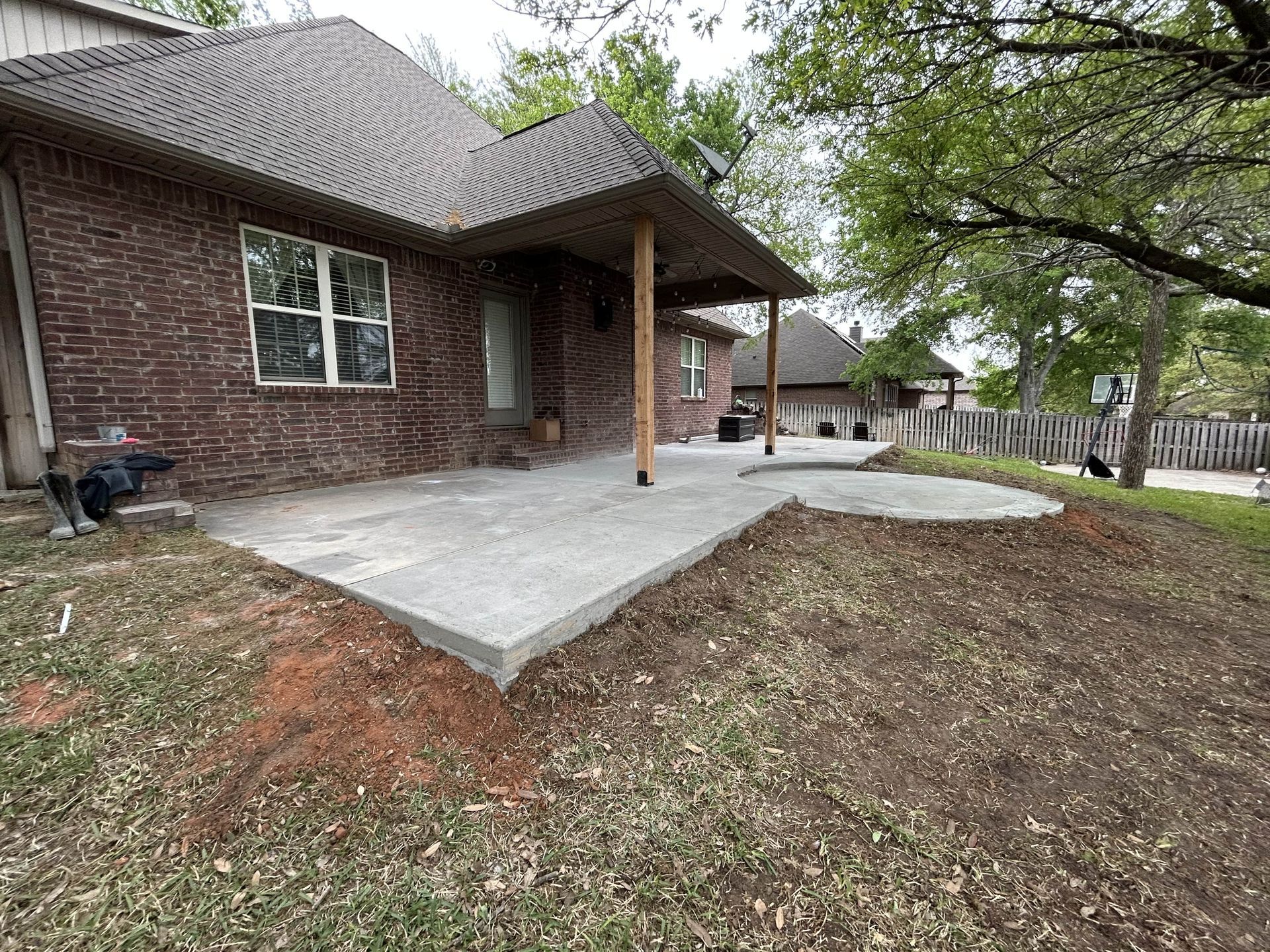 A brick house with a newly poured concrete patio extending into a backyard, bordered by dirt and trees.