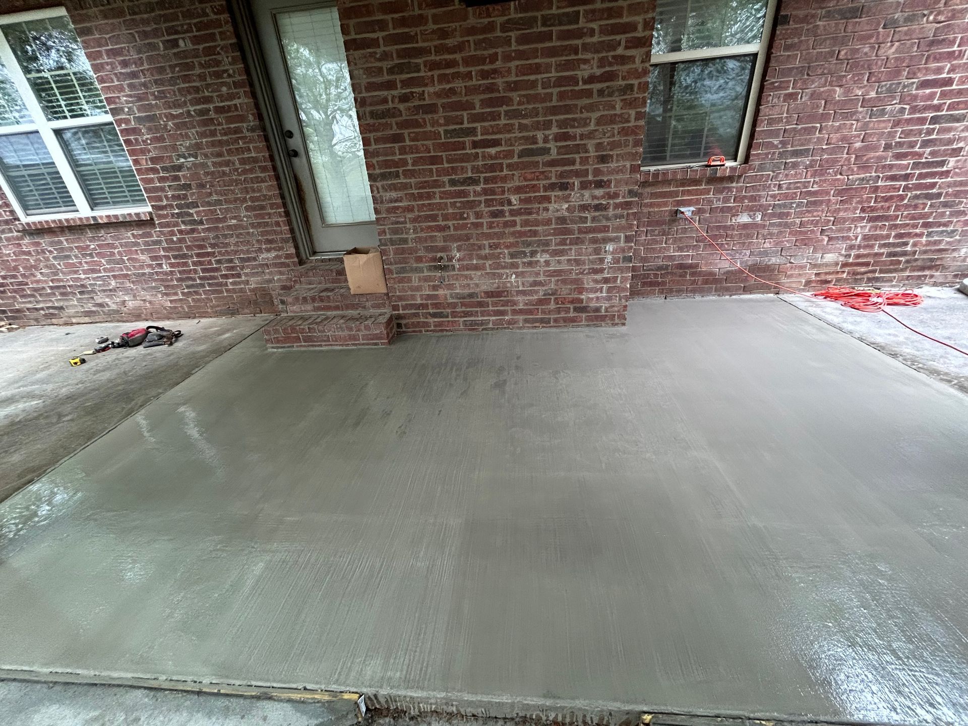 Freshly poured concrete patio extending from a brick house with two windows and a doorway.