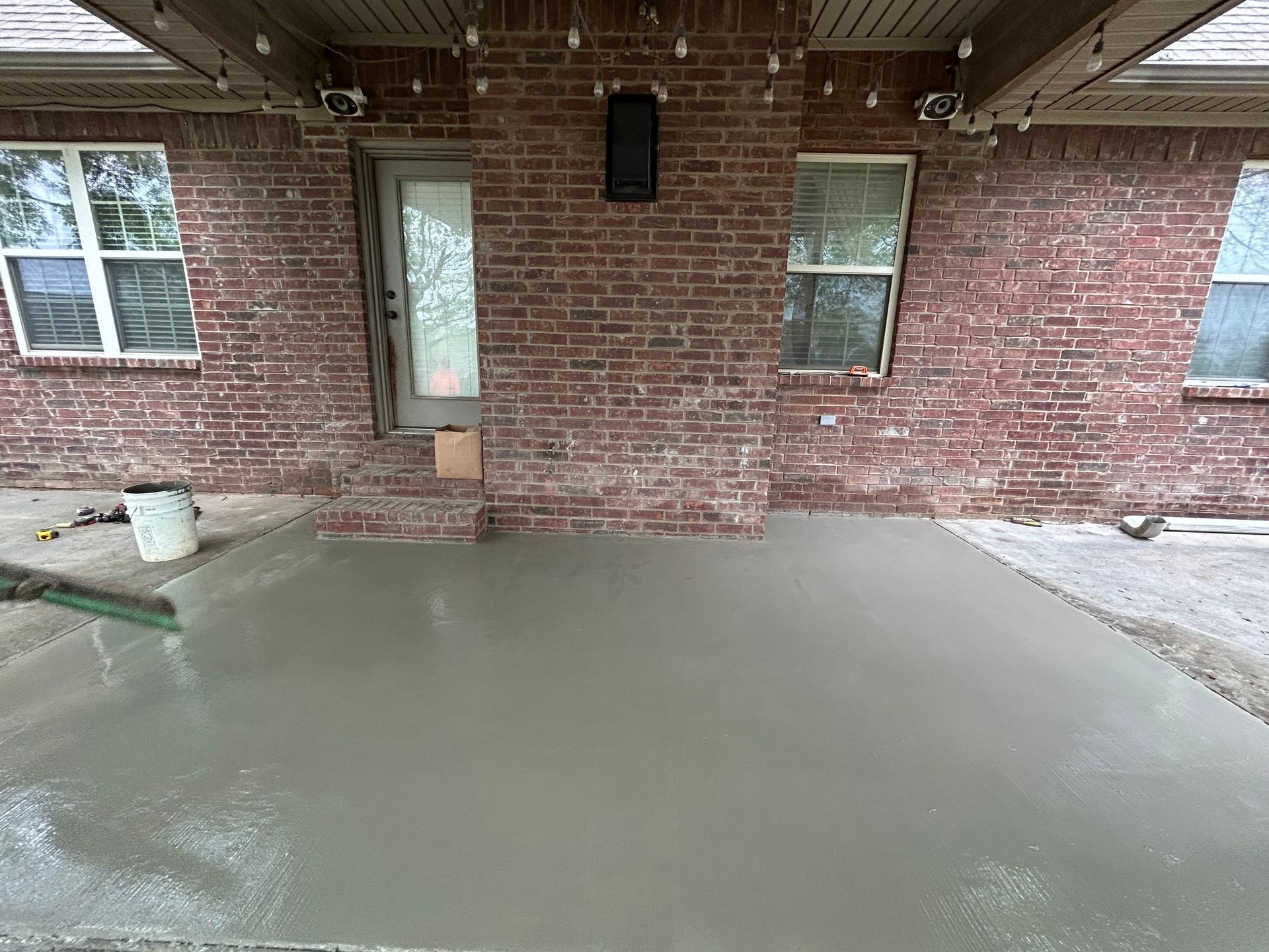 A newly poured concrete patio in front of a brick house with a door and two windows.