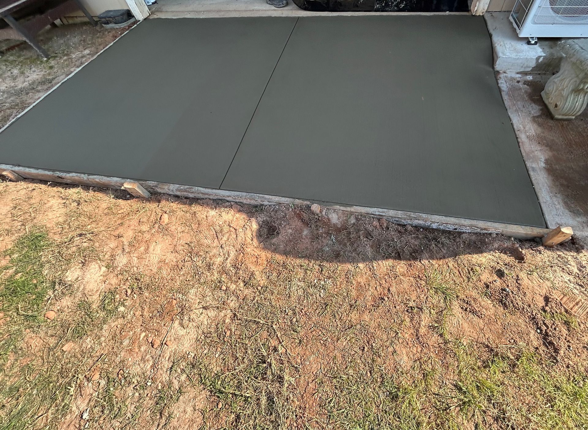 A freshly poured, gray concrete patio slab with wooden edge forms, set against a yard with grass and dirt.