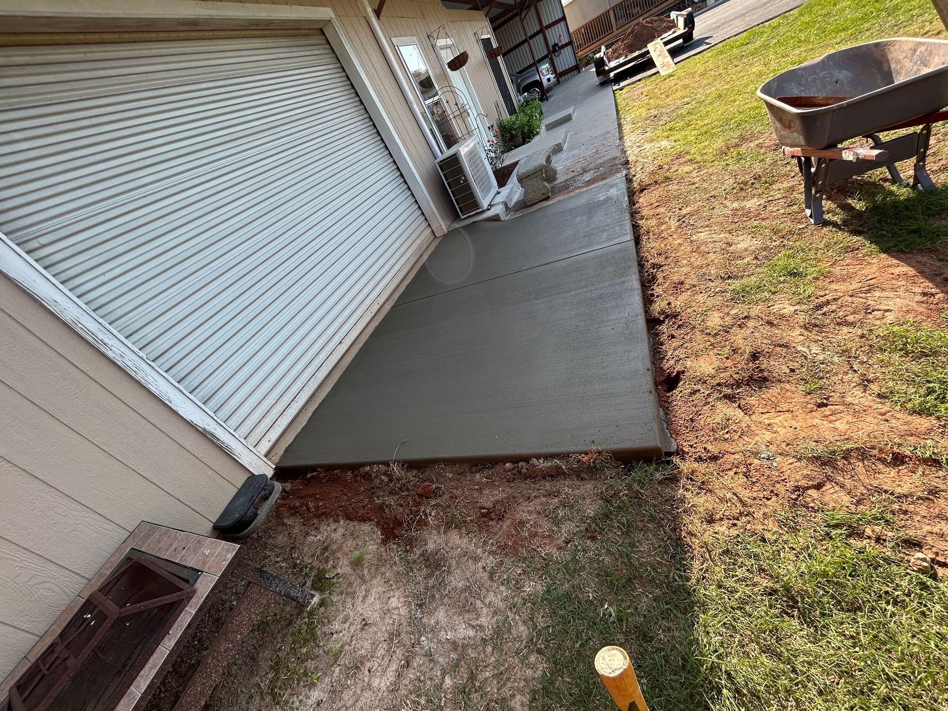 A freshly poured rectangular concrete pad sits adjacent to a garage with a white roll-up door and a dirt yard.