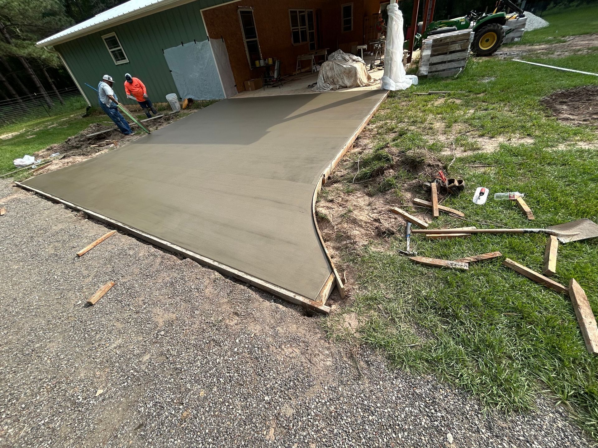 Two workers stand near a freshly poured concrete slab being installed in front of a house on a rural property.
