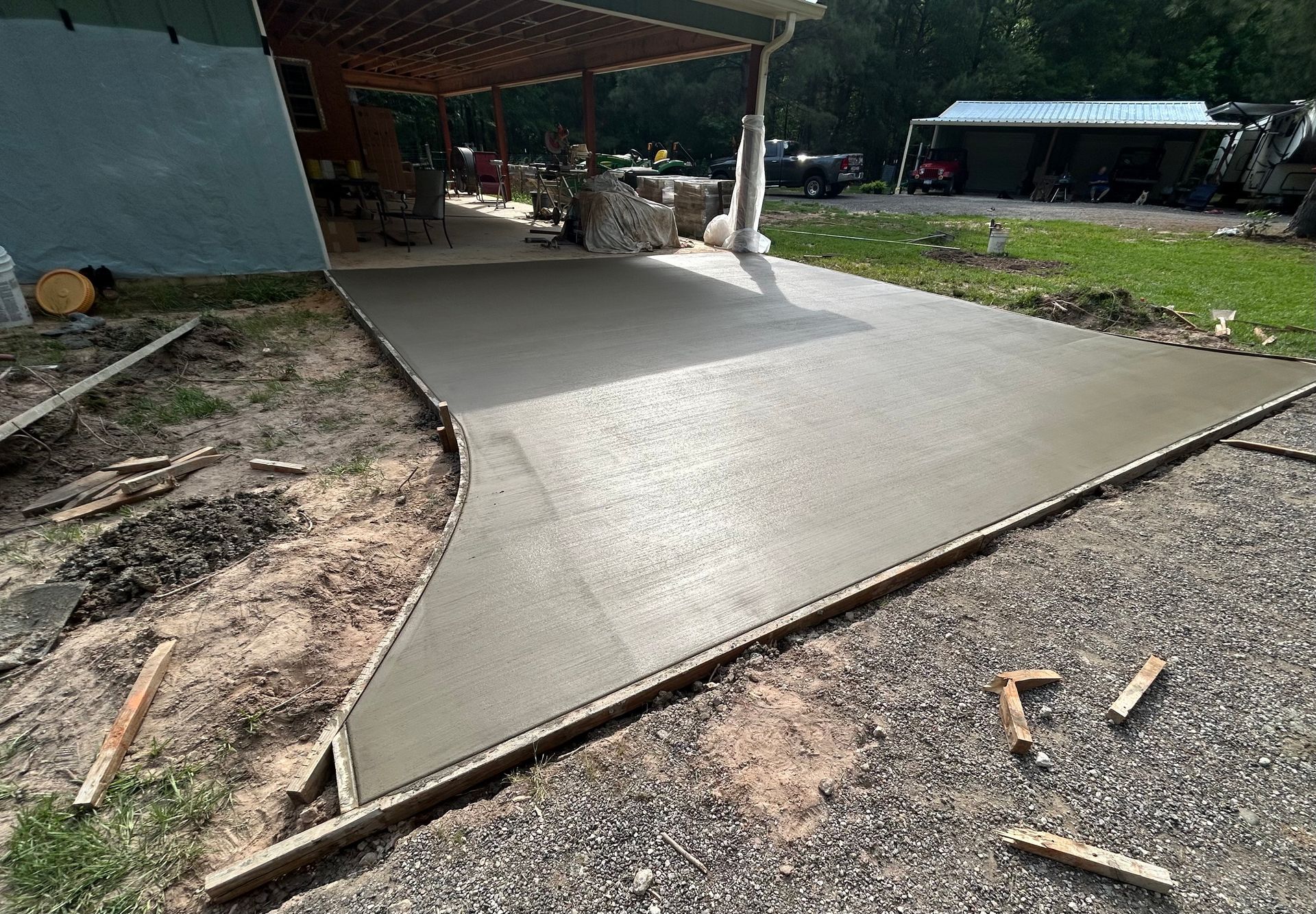A newly poured concrete driveway slab sits under a porch, bordered by gravel and dirt in a rural yard.