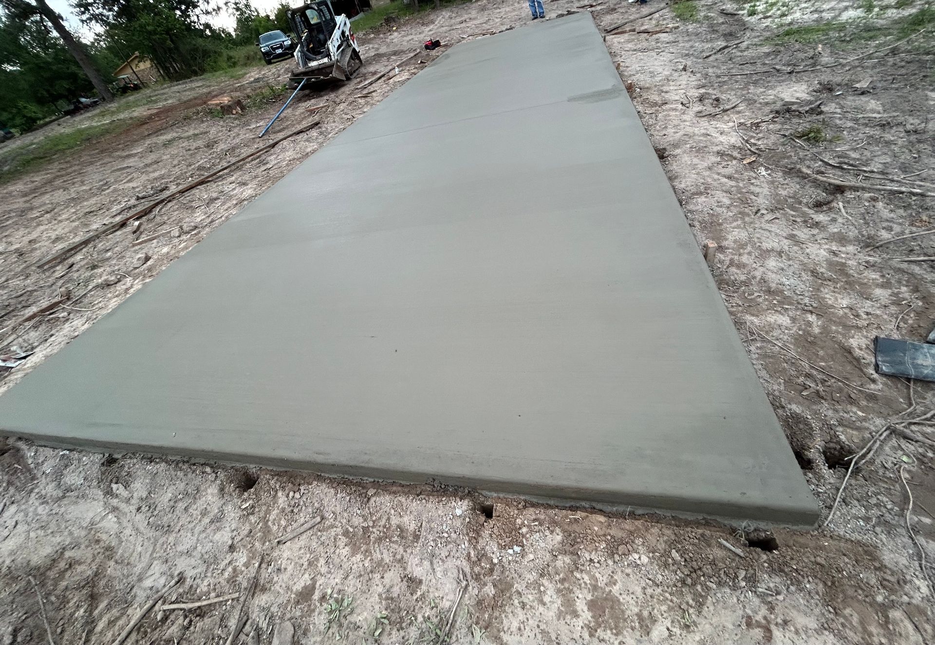 A freshly poured concrete driveway slab sits on sandy, unfinished ground next to a small construction vehicle.