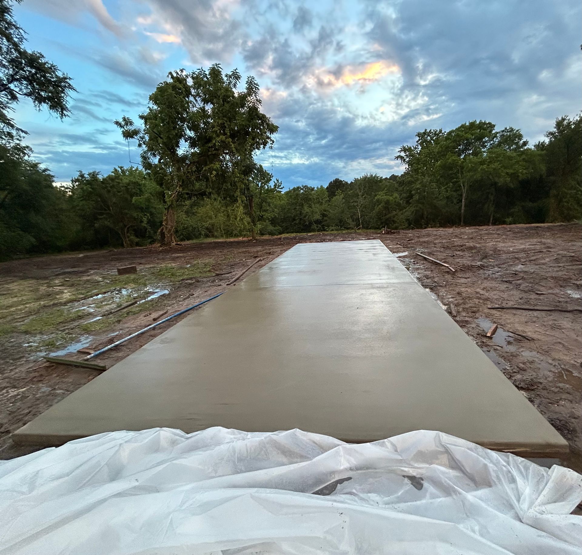 A freshly poured concrete slab in an outdoor, rural clearing under a cloudy, sunset sky.
