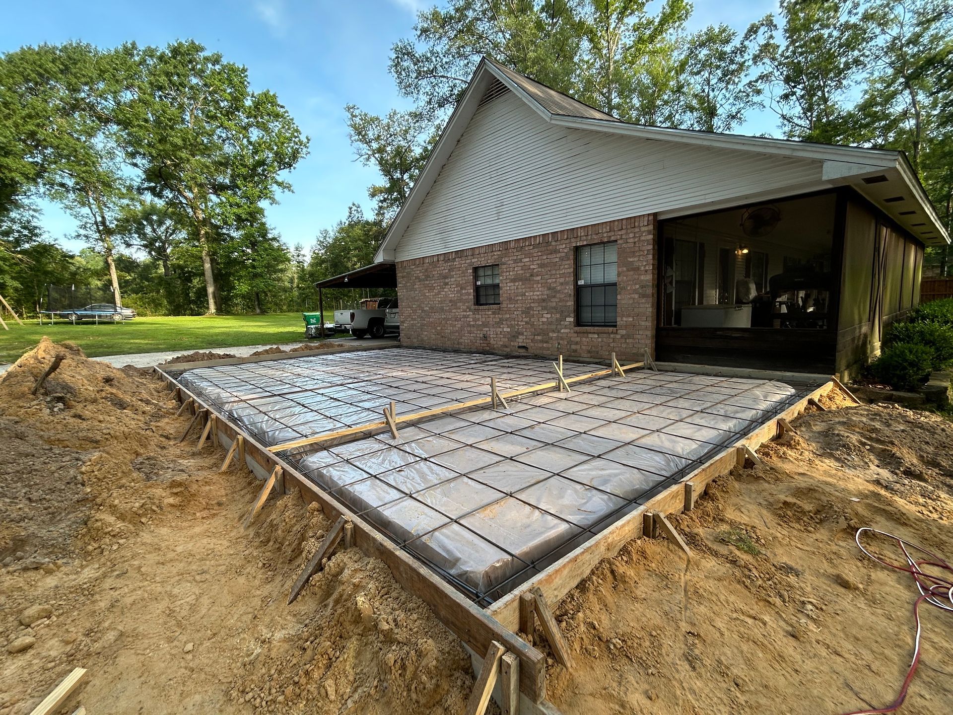 A brick house exterior under construction with a prepared concrete slab foundation reinforced with metal grid and plastic.