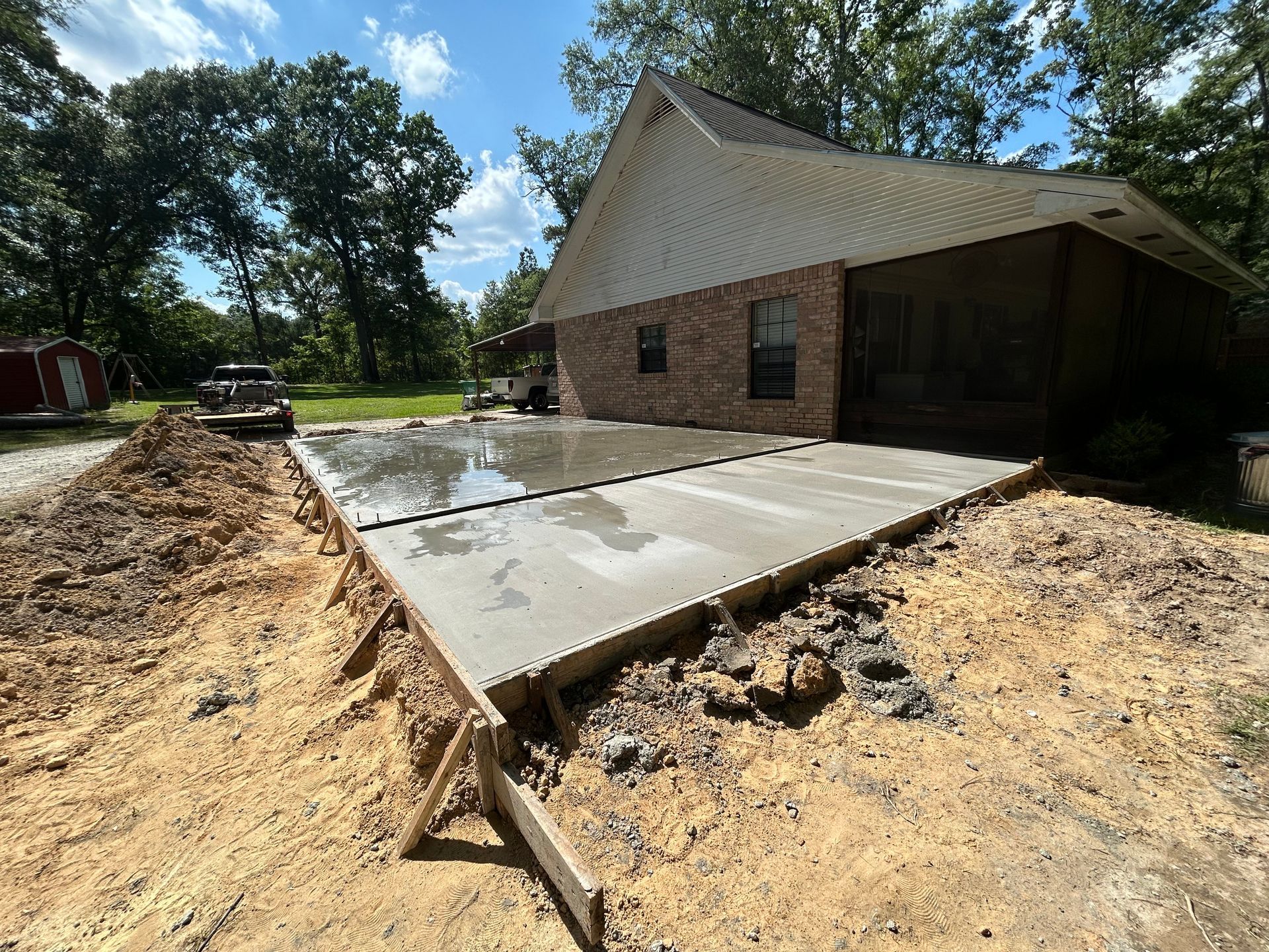 Freshly poured concrete patio extending from a brick house under construction, surrounded by dirt and a wooded yard.