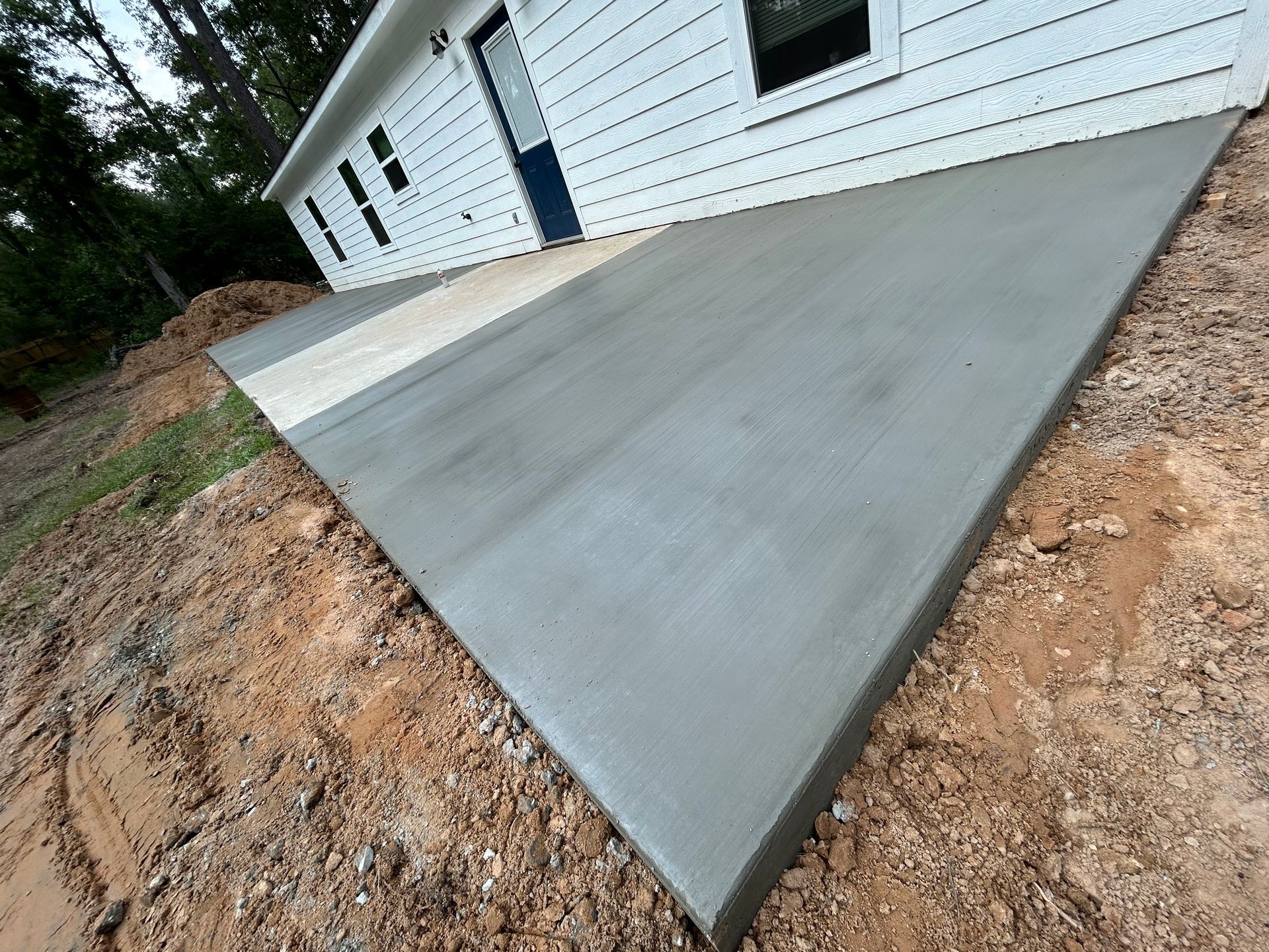 A newly poured gray concrete patio extending from the white siding of a house, surrounded by dirt and landscaping.