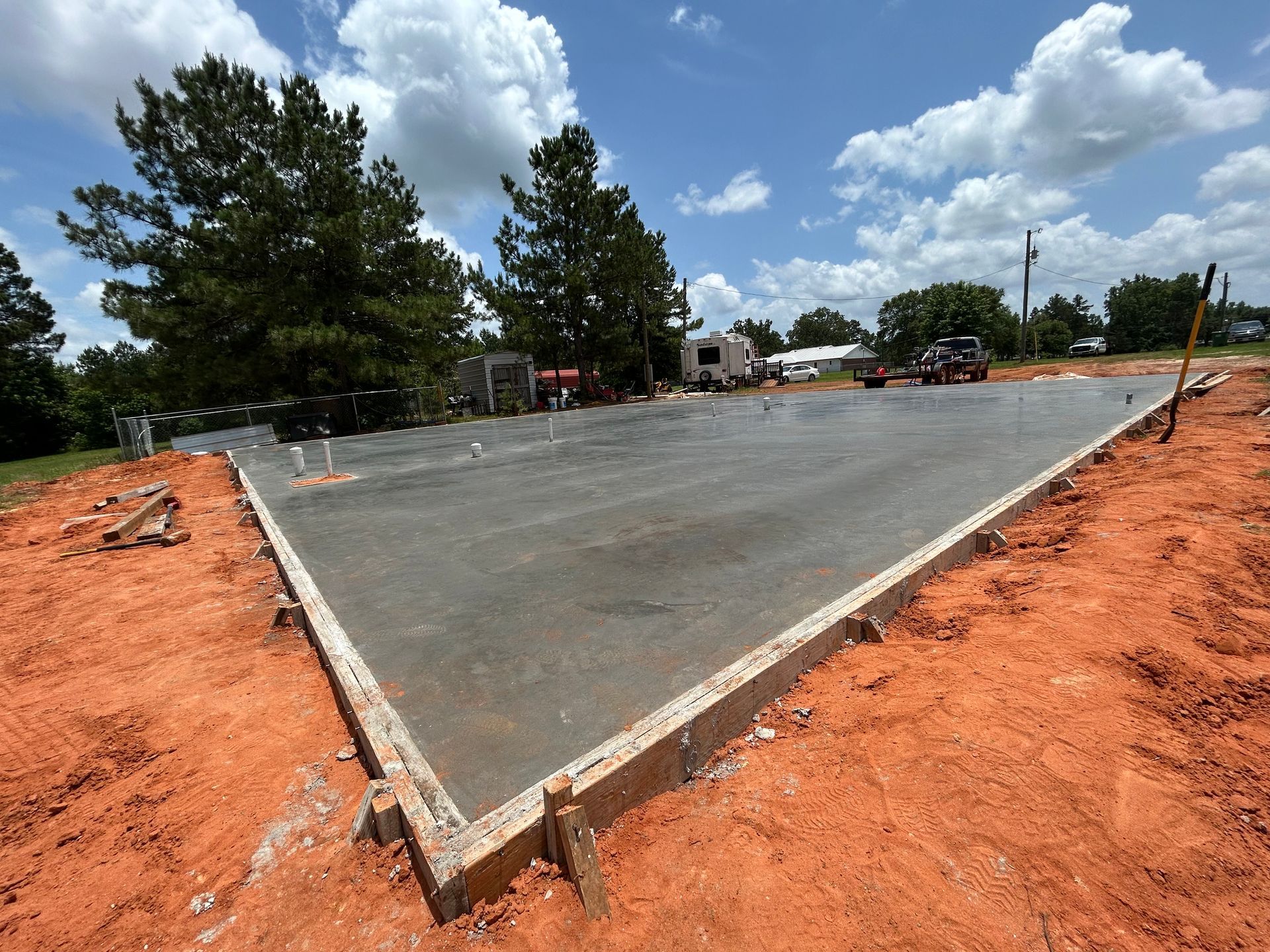 A freshly poured concrete slab foundation for a building, surrounded by red dirt under a bright, partly cloudy sky.