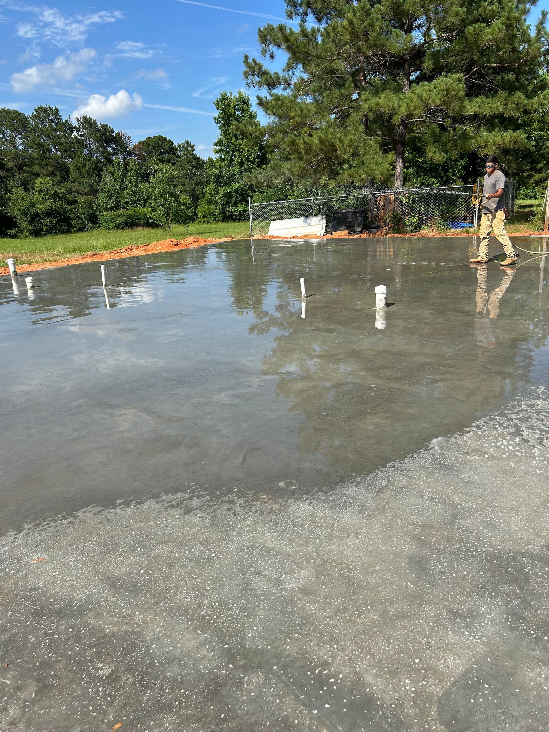 A worker walks across a freshly poured wet concrete slab foundation outdoors on a sunny day.
