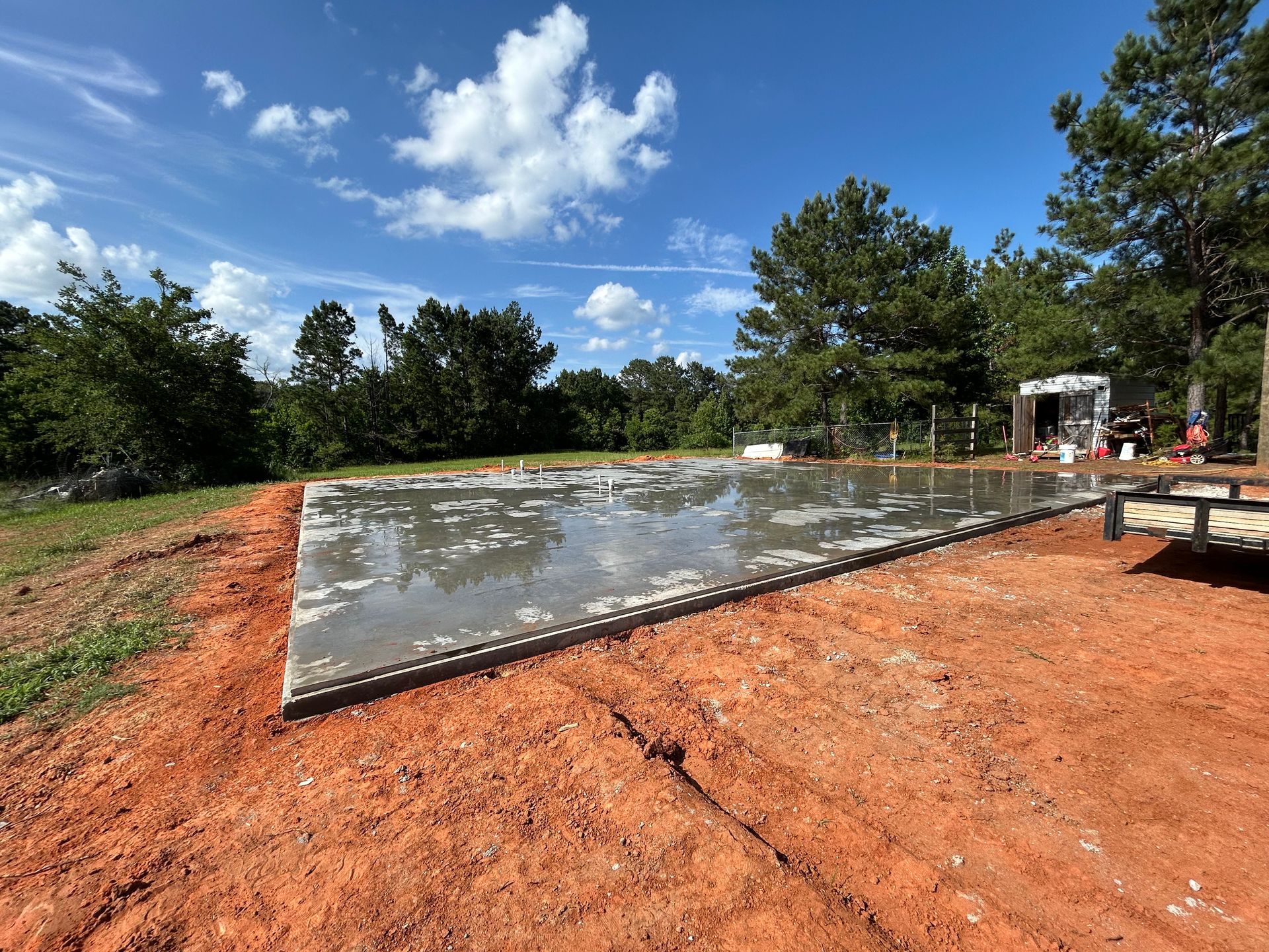 Freshly poured concrete foundation slab surrounded by red dirt on a sunny day with trees in the background.