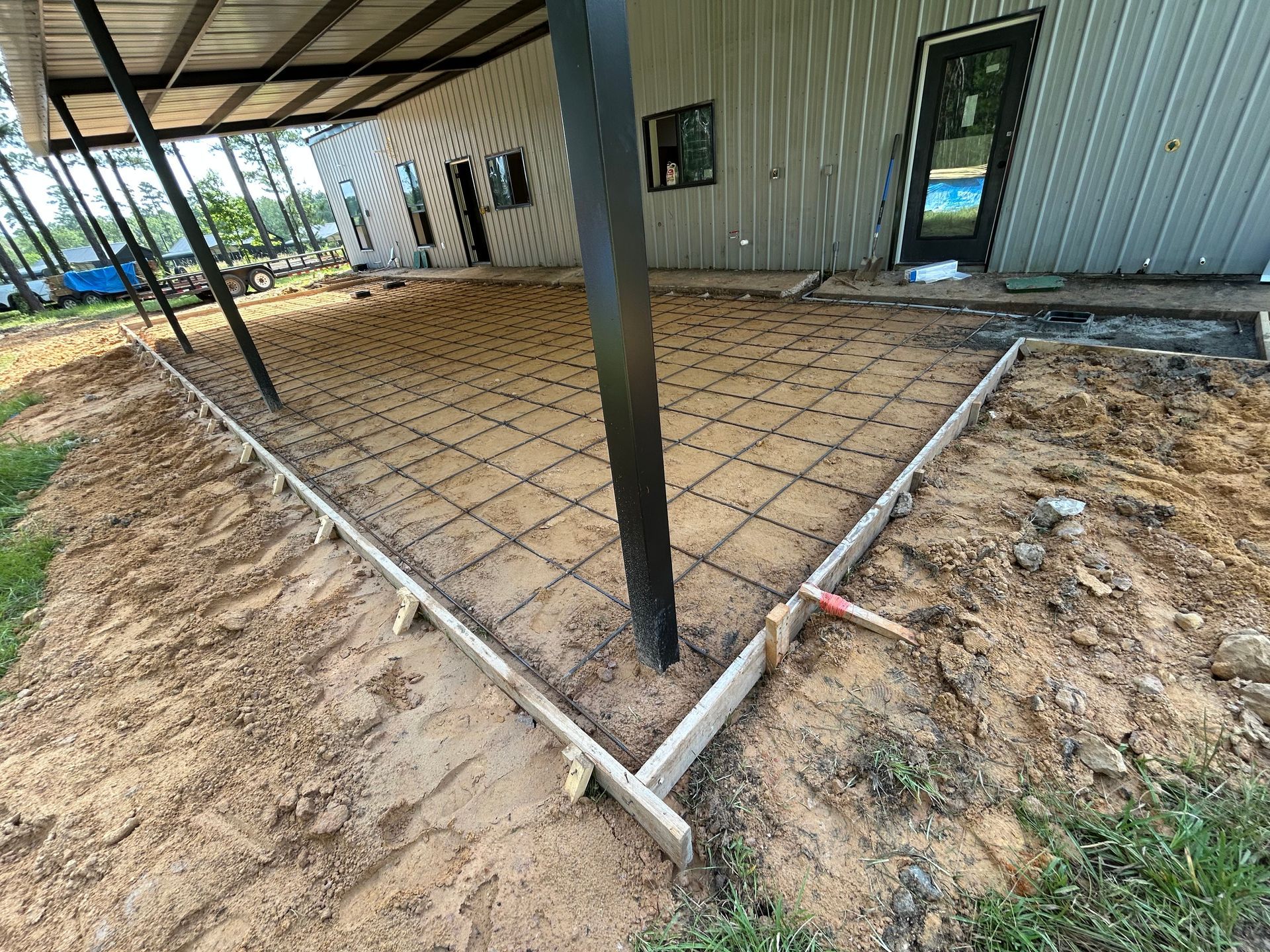 A rectangular concrete pad under construction, framed by wooden boards with wire mesh laid over sand, attached to a building.