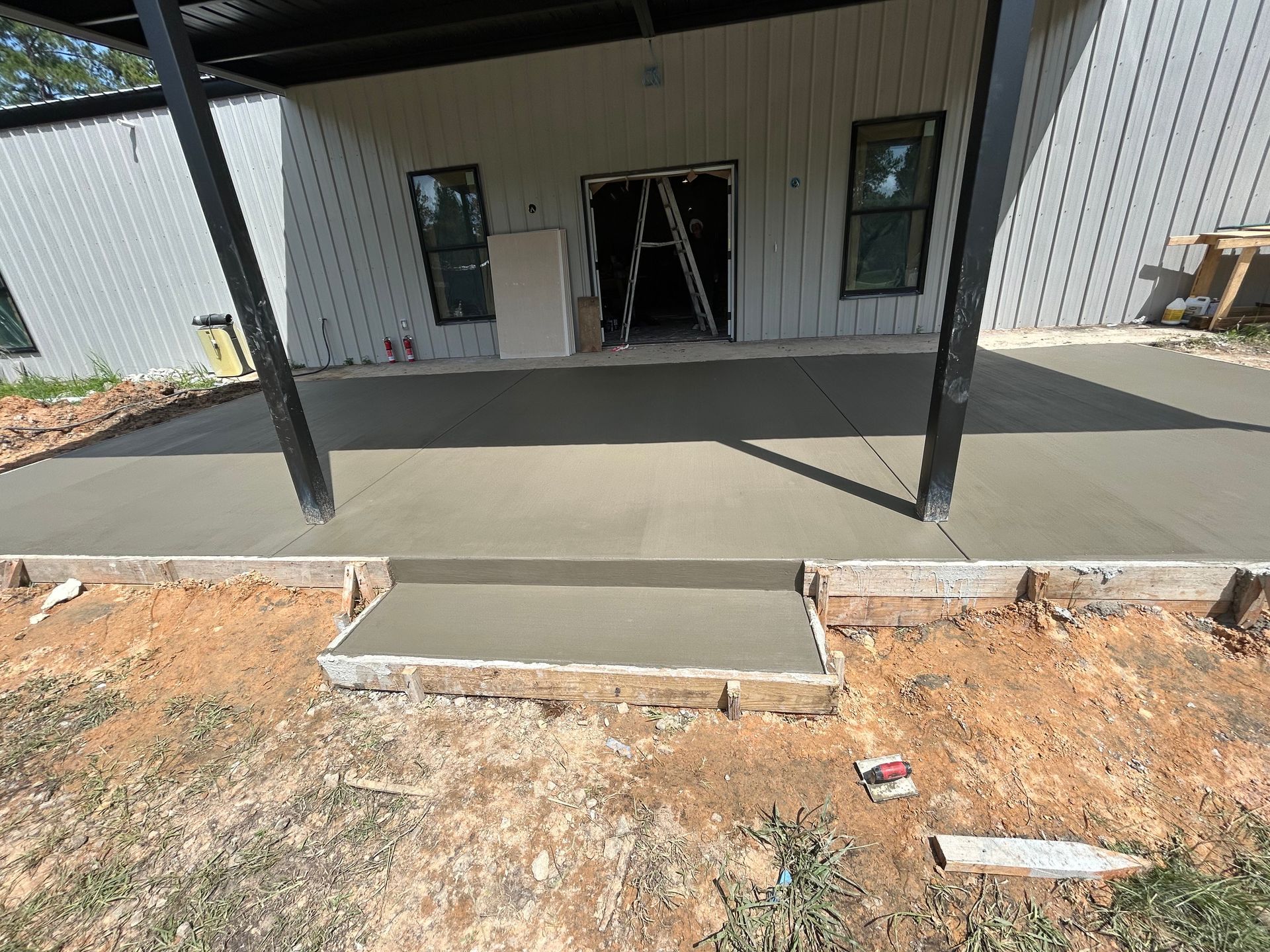 A newly poured concrete patio and step in front of a building with light gray metal siding and black structural supports.