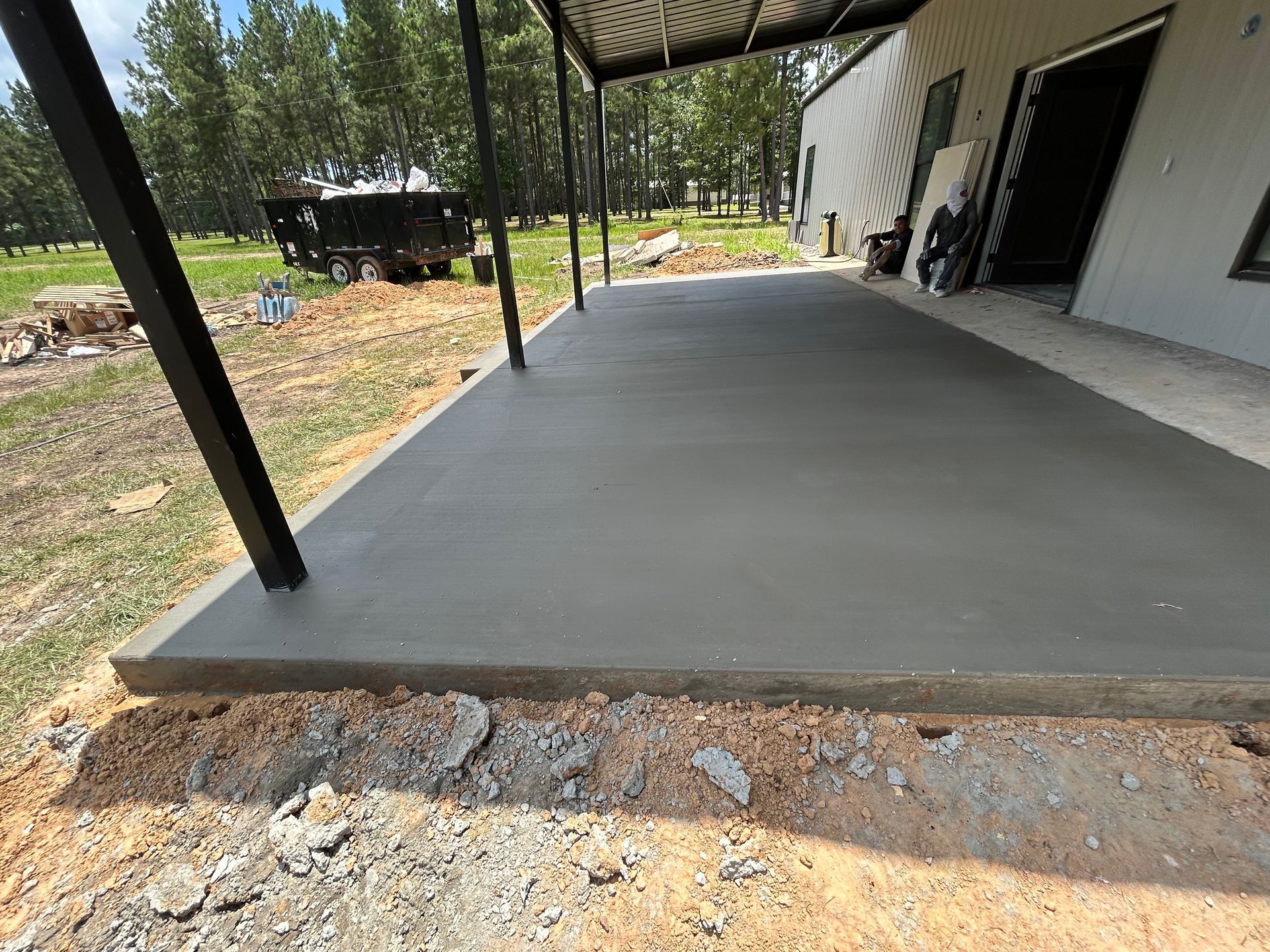 Freshly poured concrete patio extending from a building with black metal support posts, set against a grassy landscape.