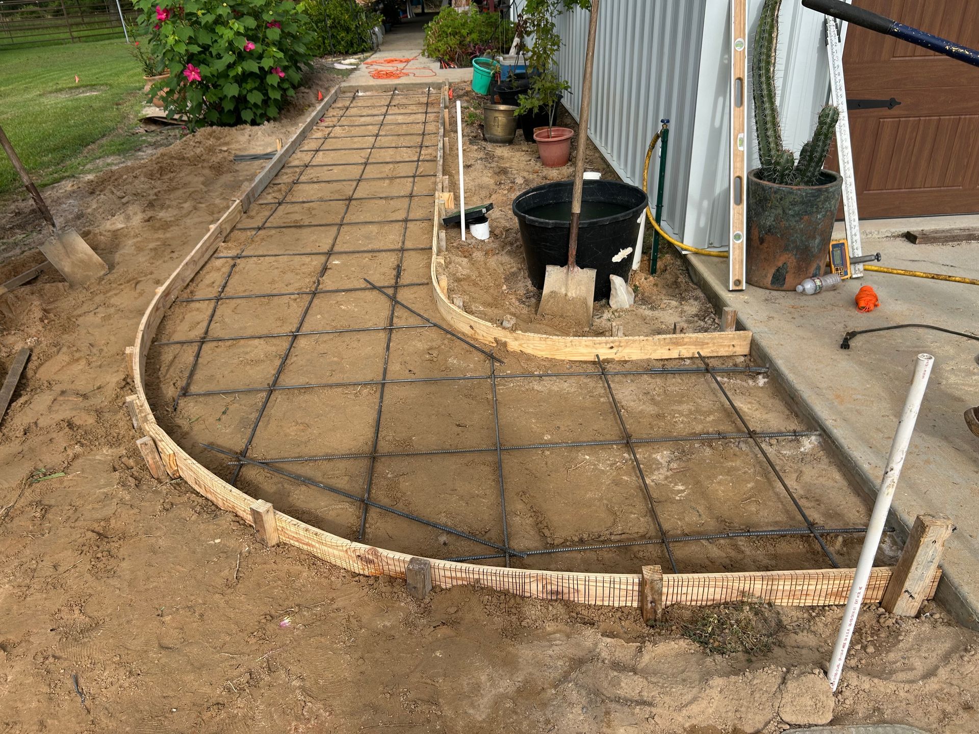 A curved concrete walkway under construction with wooden forms and metal reinforcement grid on a sandy surface.