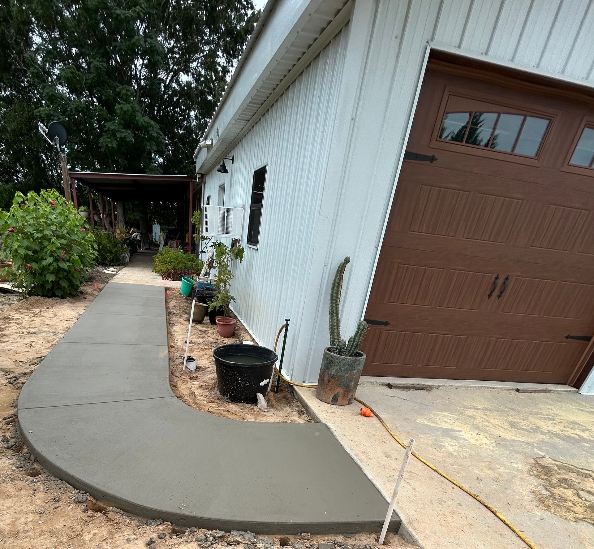 A newly poured gray concrete walkway curves alongside a building with white siding and a brown garage door.