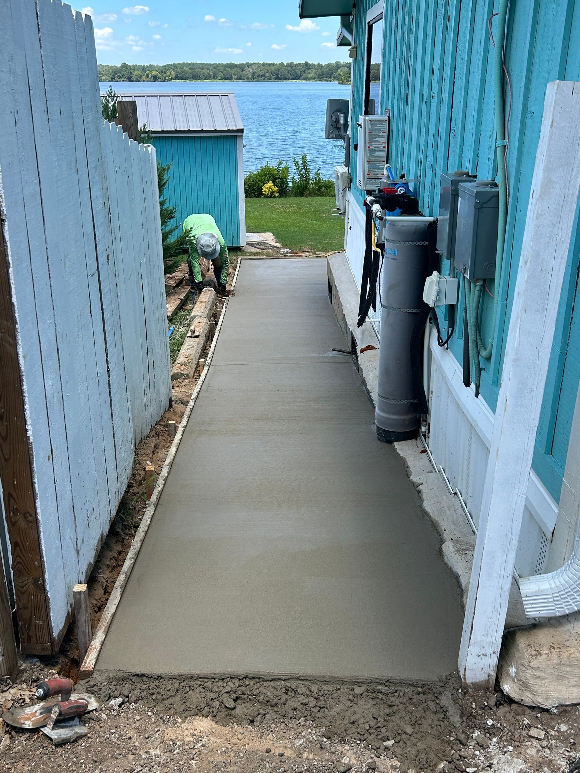 A freshly poured concrete sidewalk between a blue mobile home and a white lattice fence, leading toward a lake.