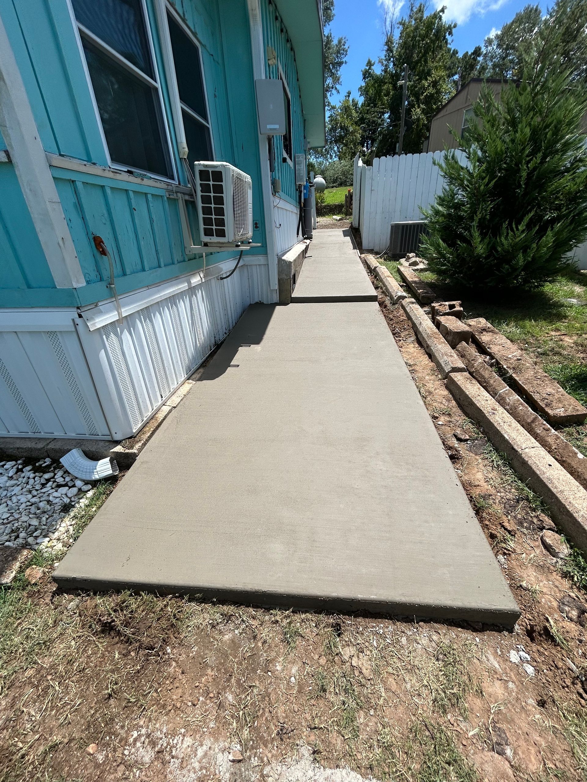 A new concrete walkway runs alongside a teal mobile home, bordered by a wooden edge on the right.