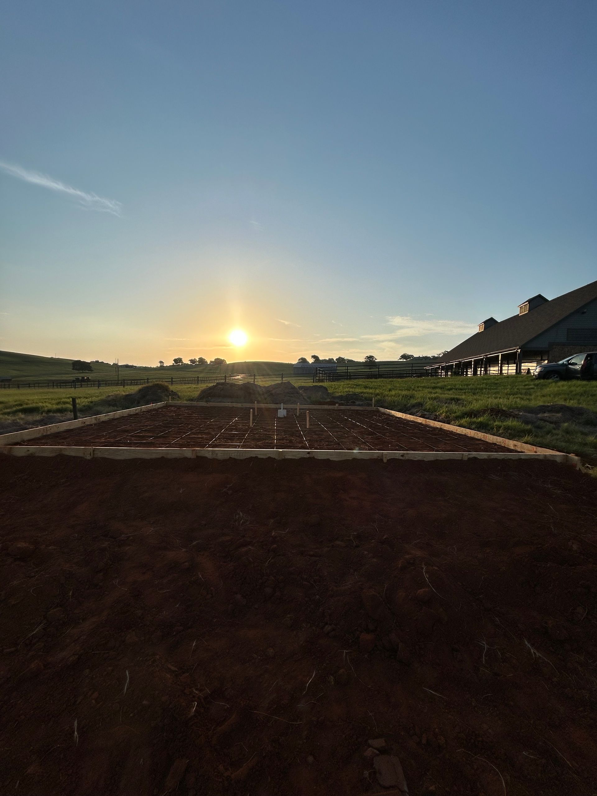 A rectangular foundation framed with wood boards set in a dirt field under a bright sunset.