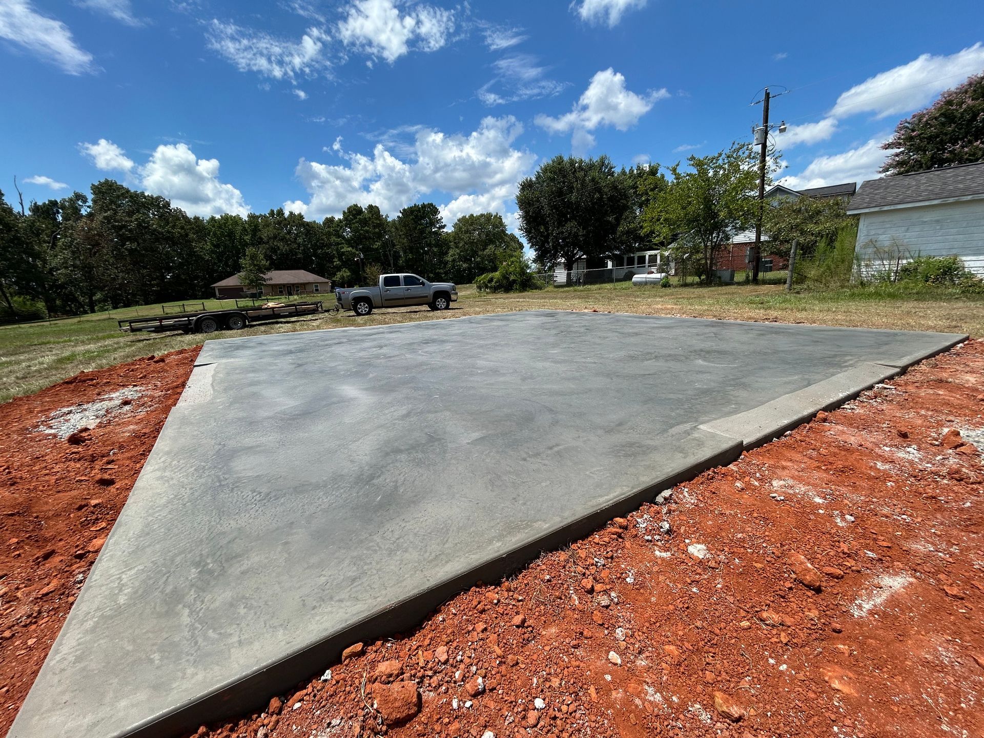 A freshly poured concrete slab sits in a yard with red soil, surrounded by trees and a distant house under a blue sky.