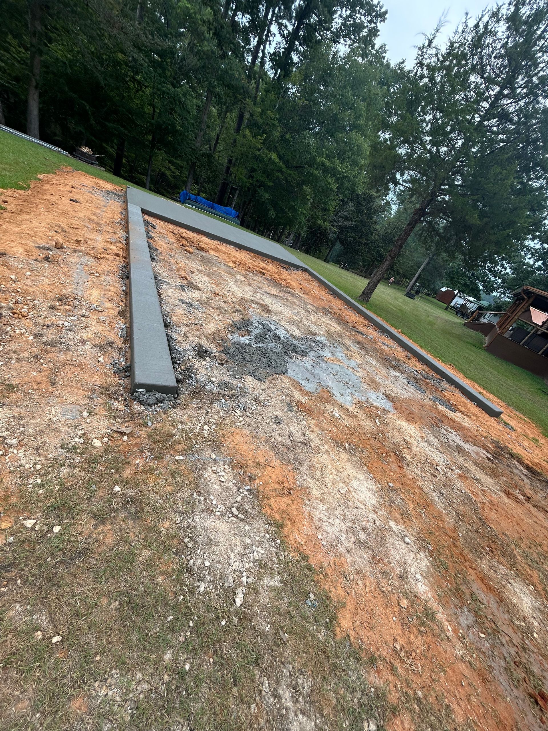 A concrete border outlining a rectangular area in a grassy, wooded yard filled with wood chips and gravel.