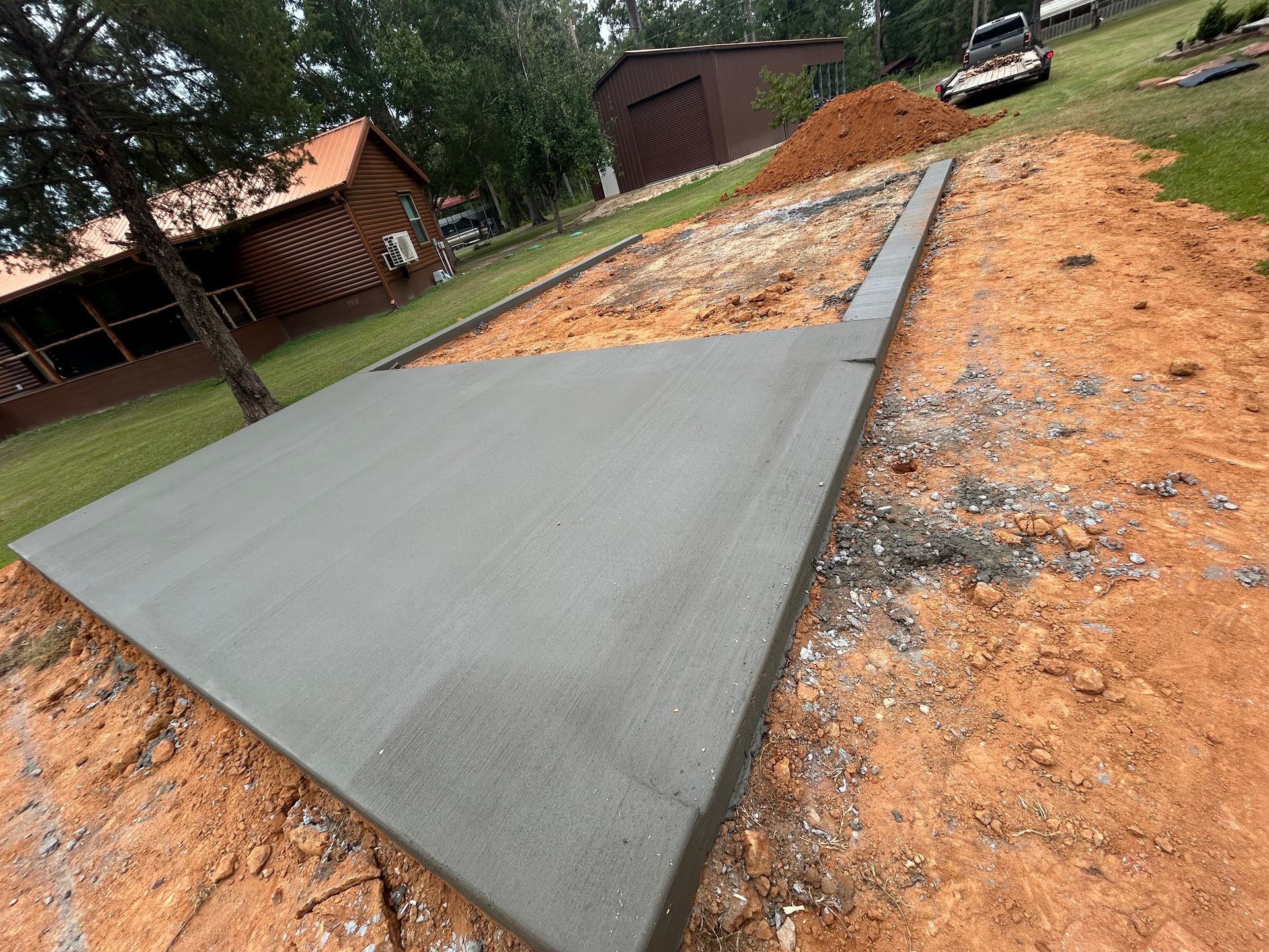 A newly poured concrete slab sits in an outdoor area of reddish dirt, with a wooden barn in the background.