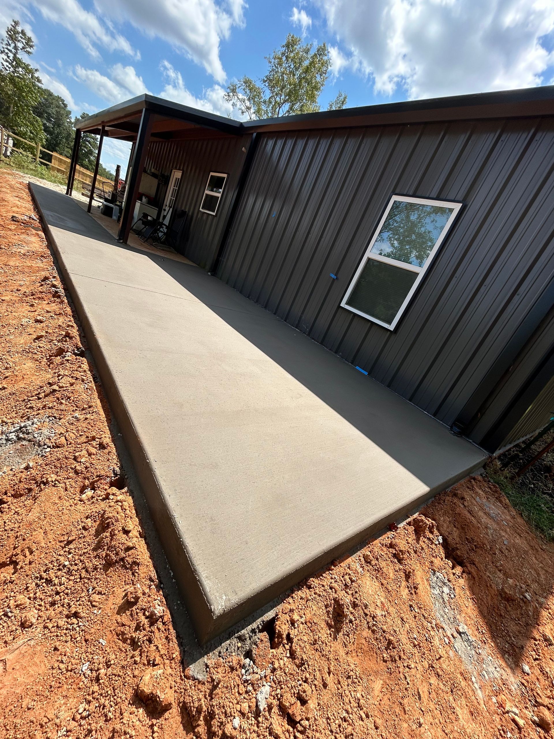 A new gray concrete porch wraps around the side of a modern dark metal building under a blue sky with scattered clouds.