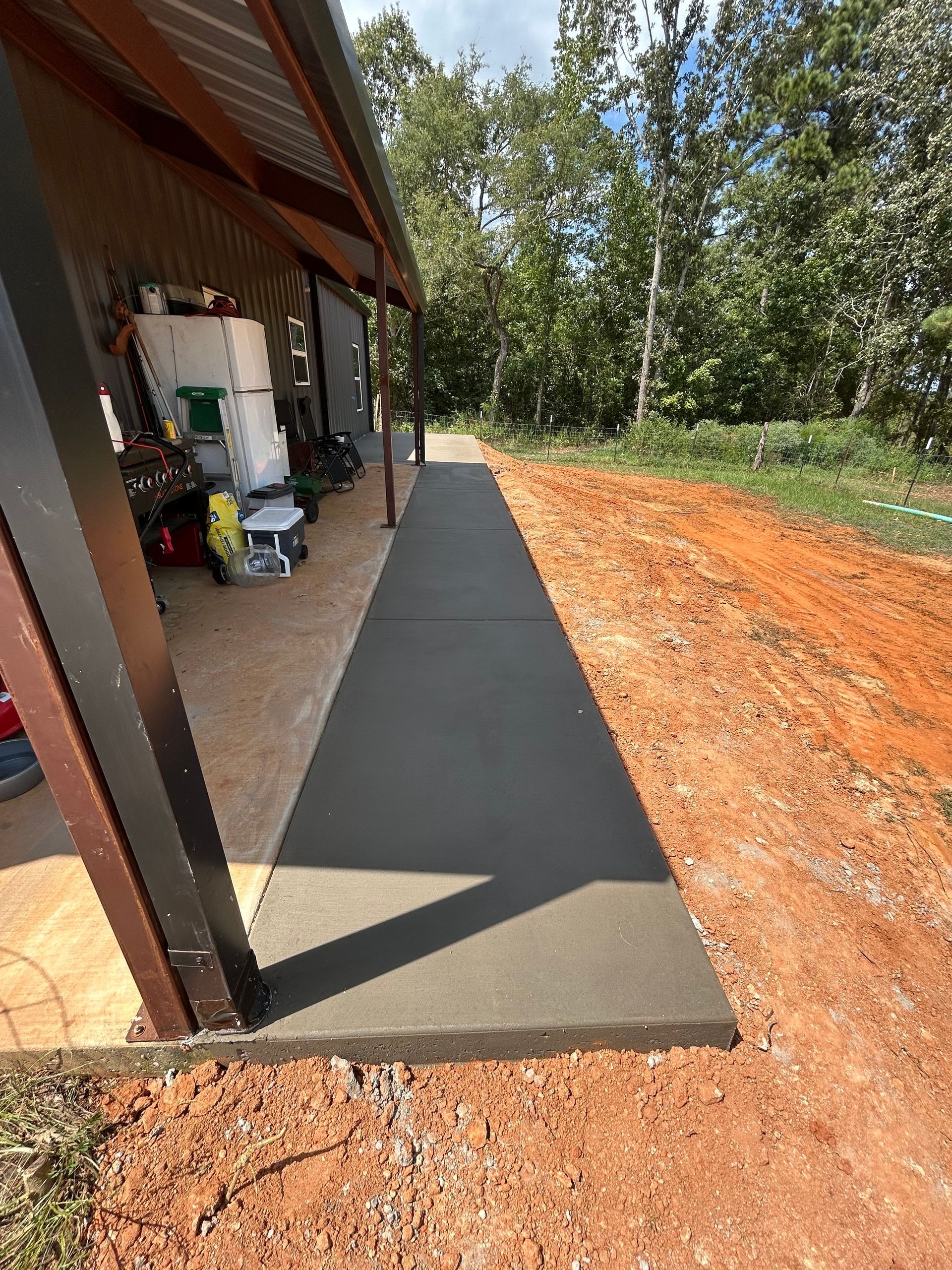 A newly poured concrete walkway runs along the side of a metal-sided building next to a dirt embankment and trees.