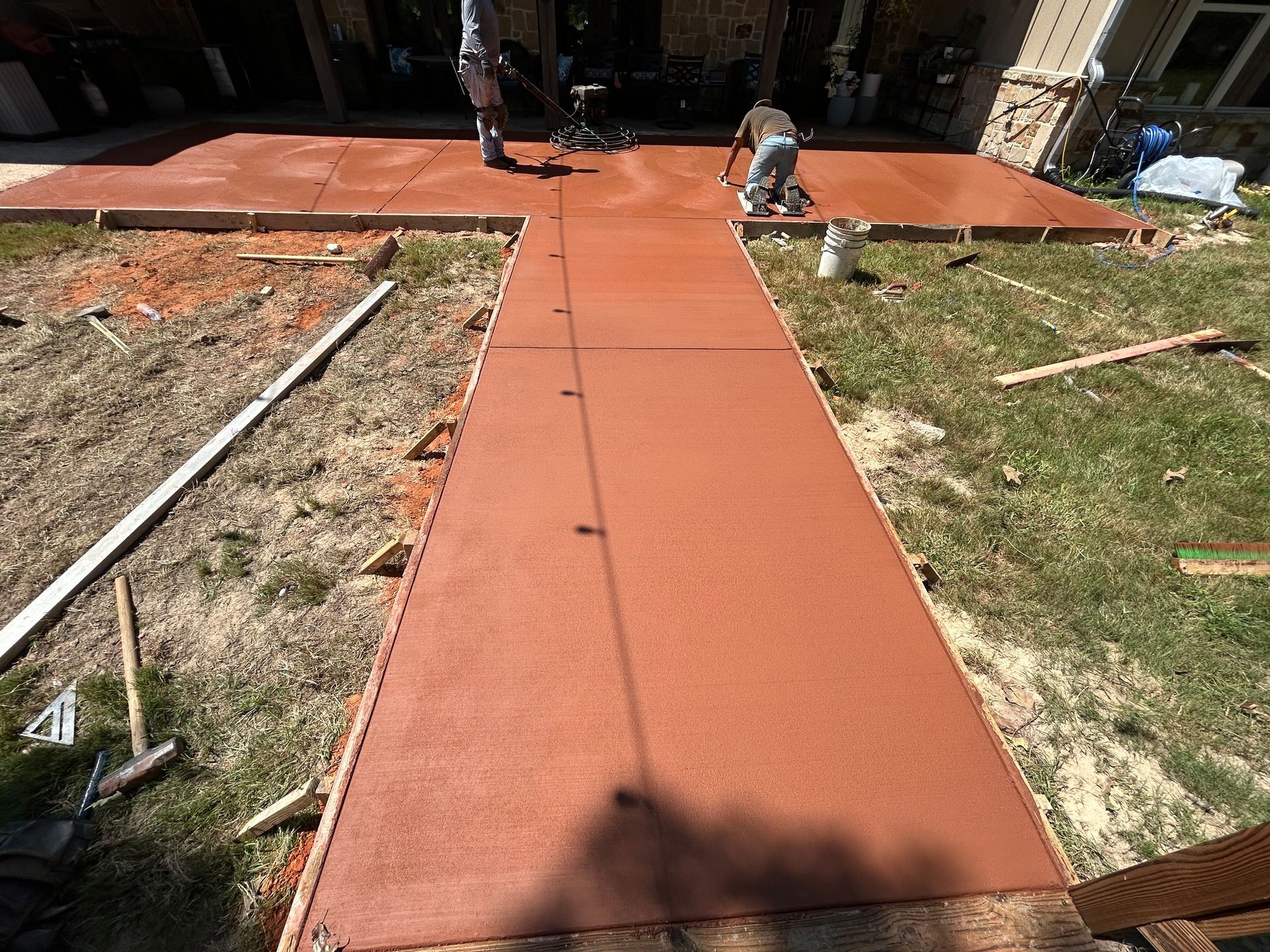 Two workers smooth freshly poured, reddish-brown concrete for a new walkway and patio area in a residential backyard.