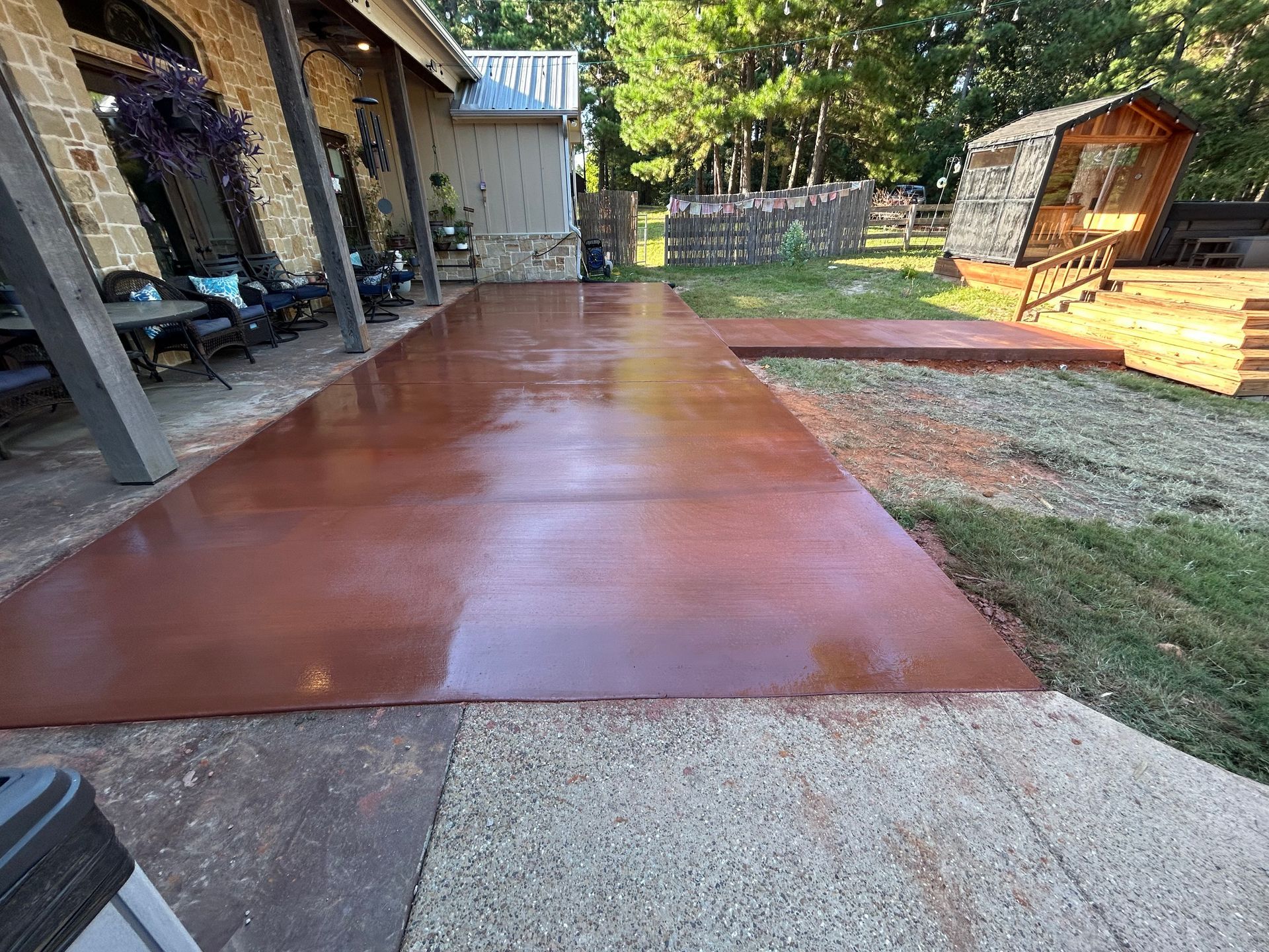 A freshly stained, glossy reddish-brown concrete patio extends from a stone house toward a grassy yard and small structure.