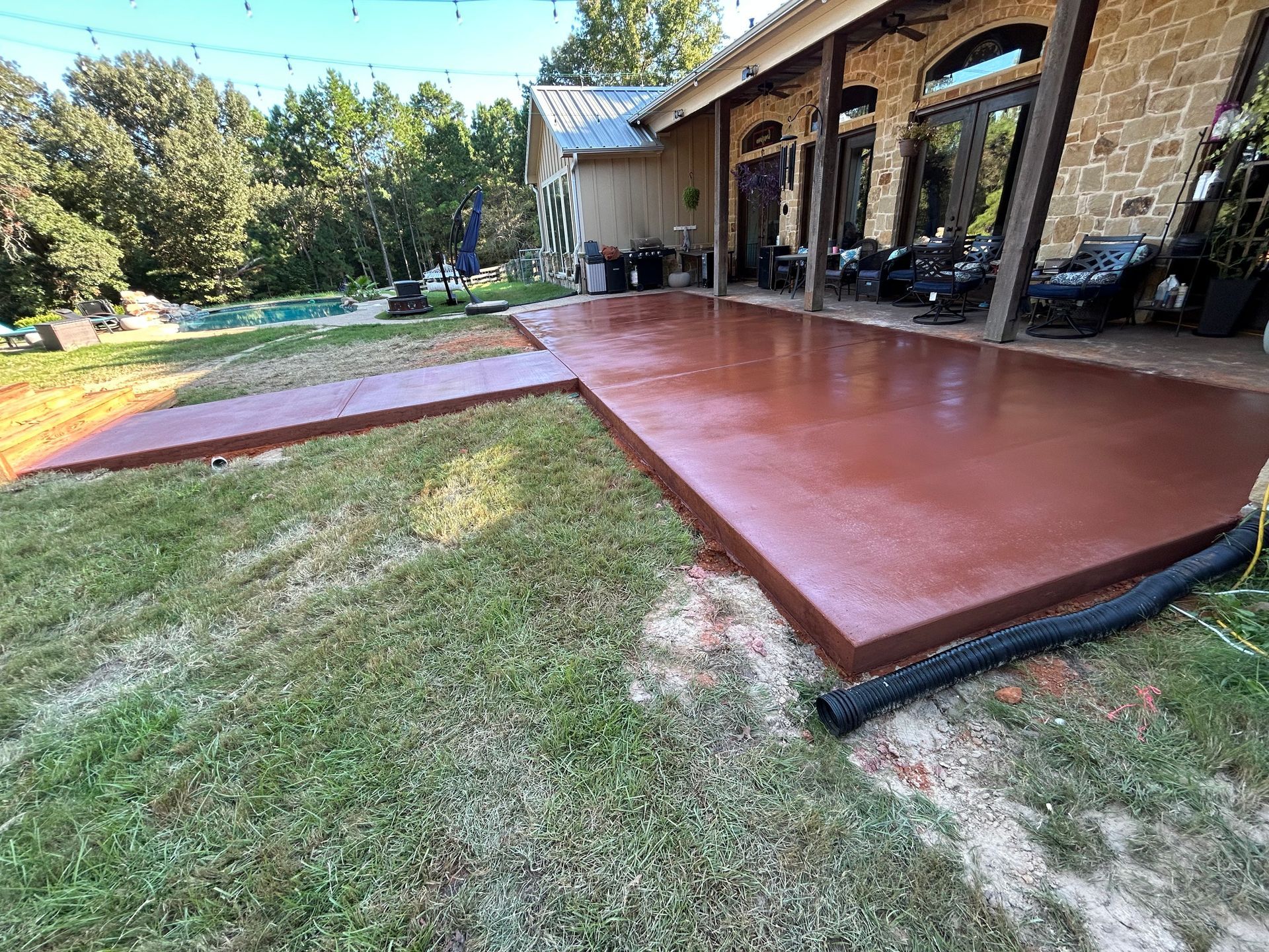 A newly installed, reddish-brown concrete patio and walkway extend from a stone-walled house into a grassy backyard.