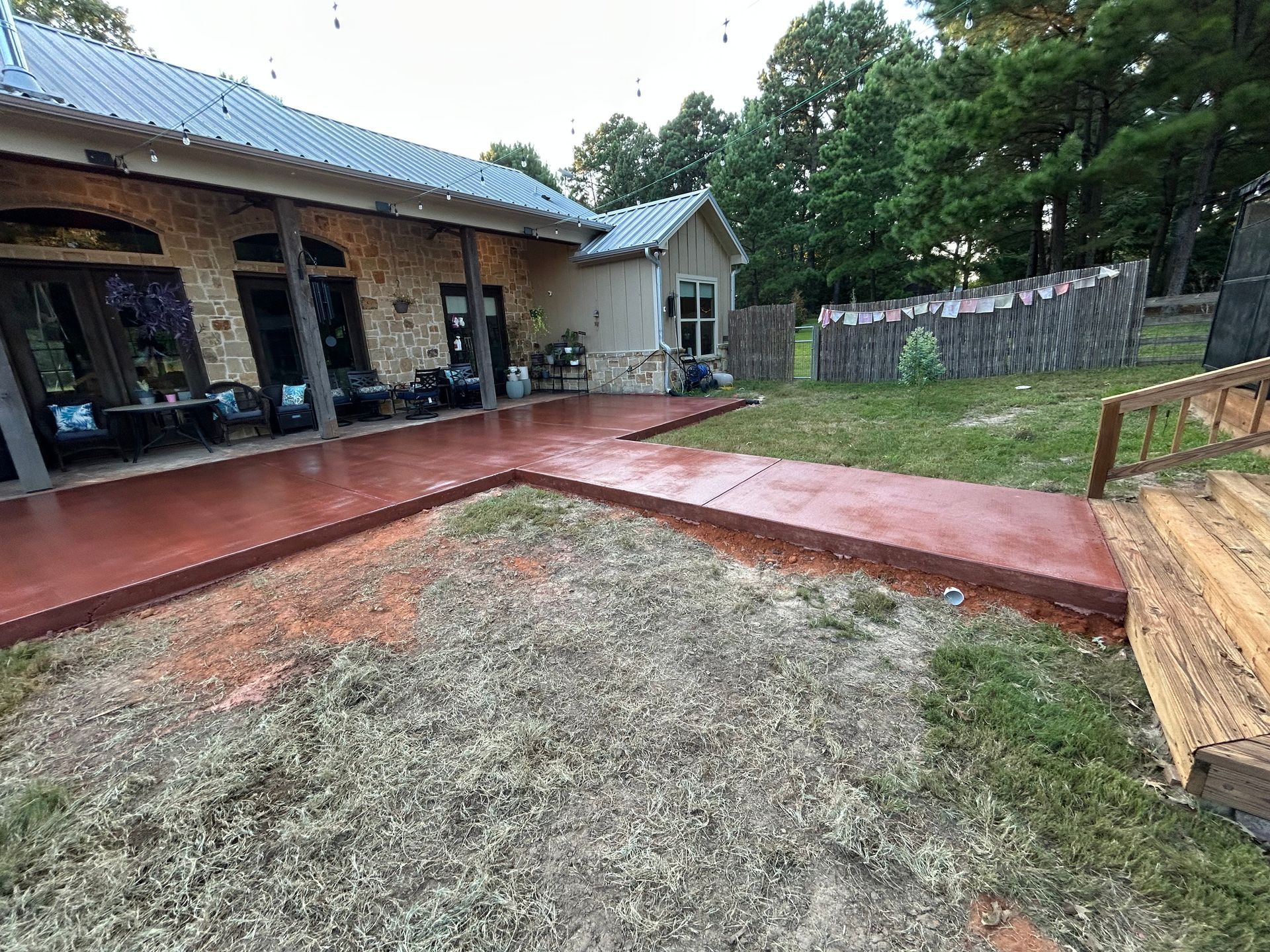A stone house with a red-painted concrete patio and walkway leading to wooden stairs in a grassy backyard.