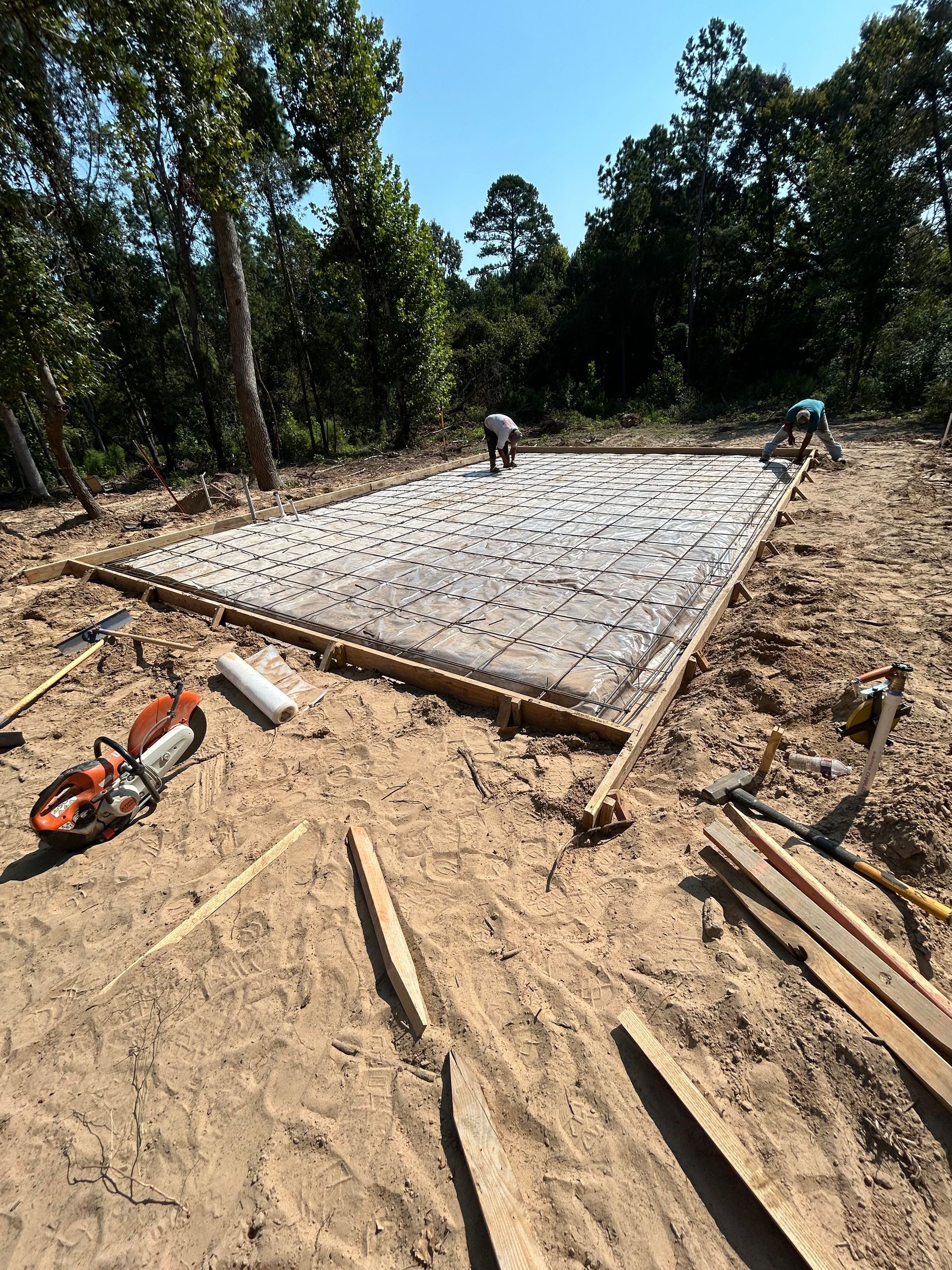 Workers prepare a concrete foundation slab for construction, surrounded by wooden forms and equipment on a dirt site.