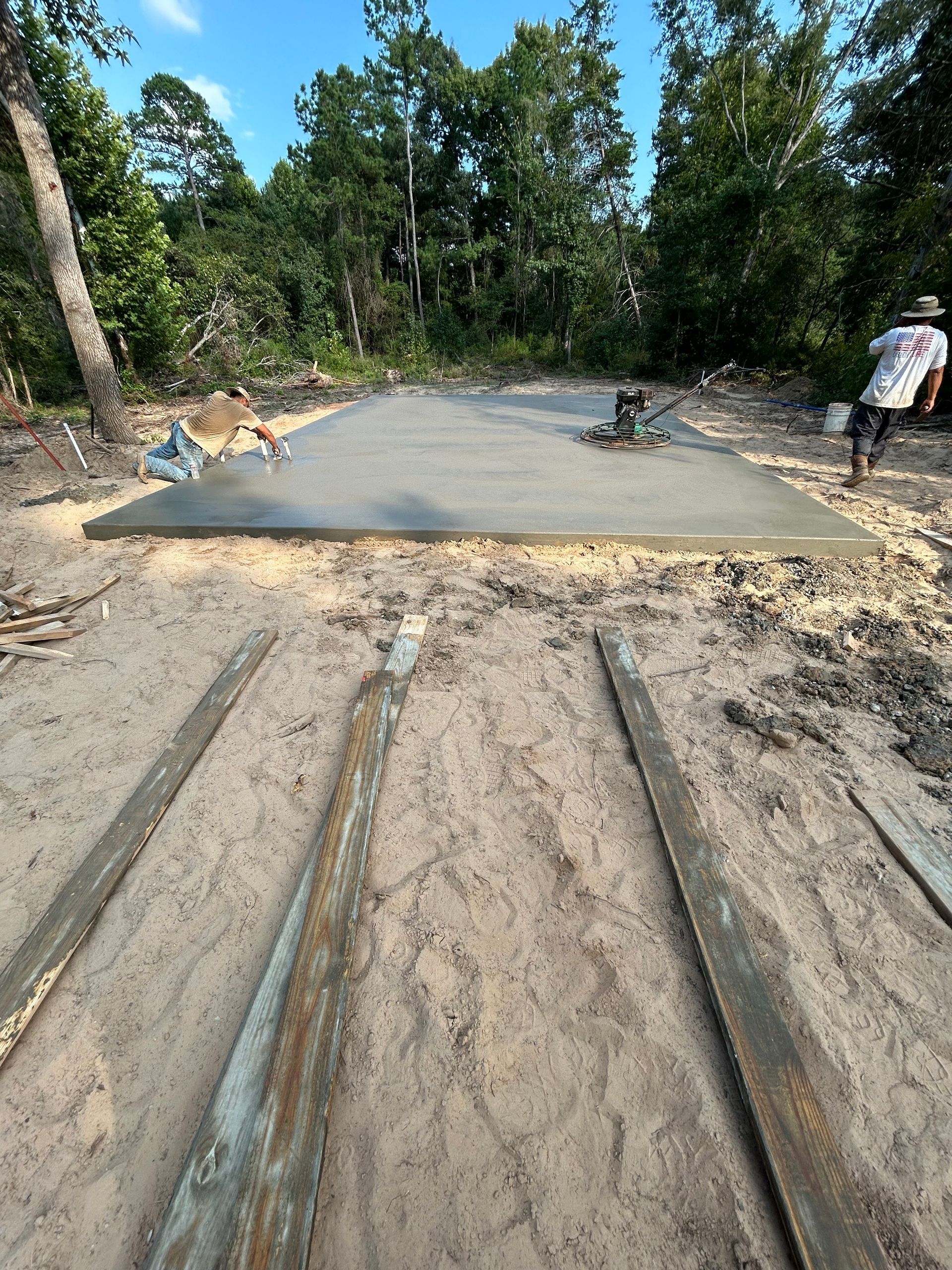 A person works on a freshly poured rectangular concrete slab in a wooded, sandy construction site.