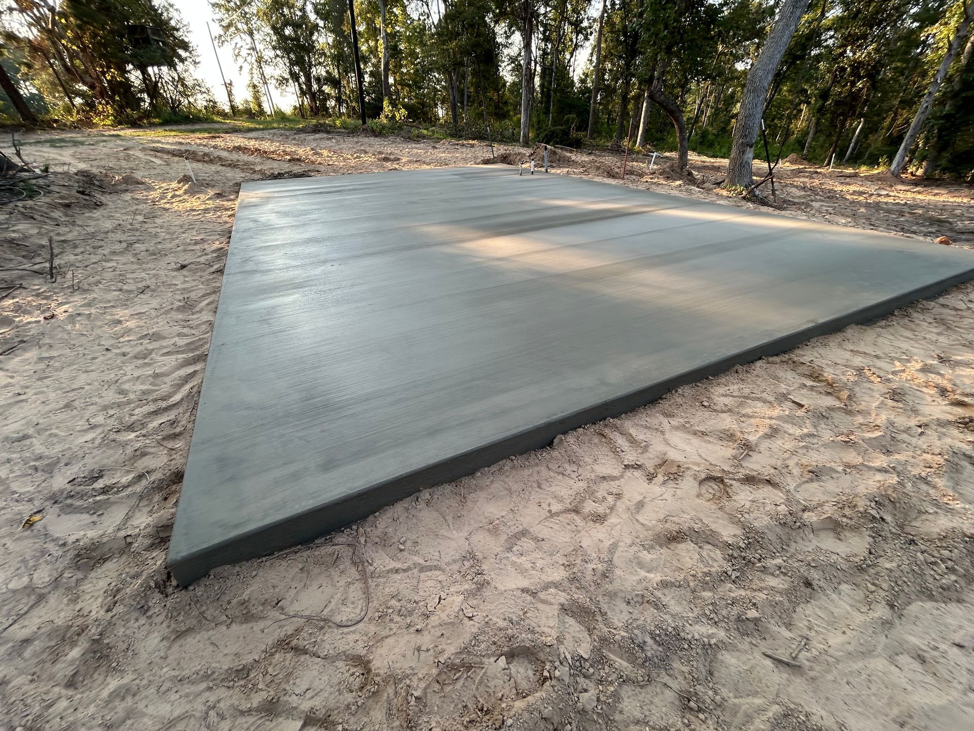 A freshly poured, smooth rectangular concrete slab sits on sandy ground surrounded by trees in a wooded area.