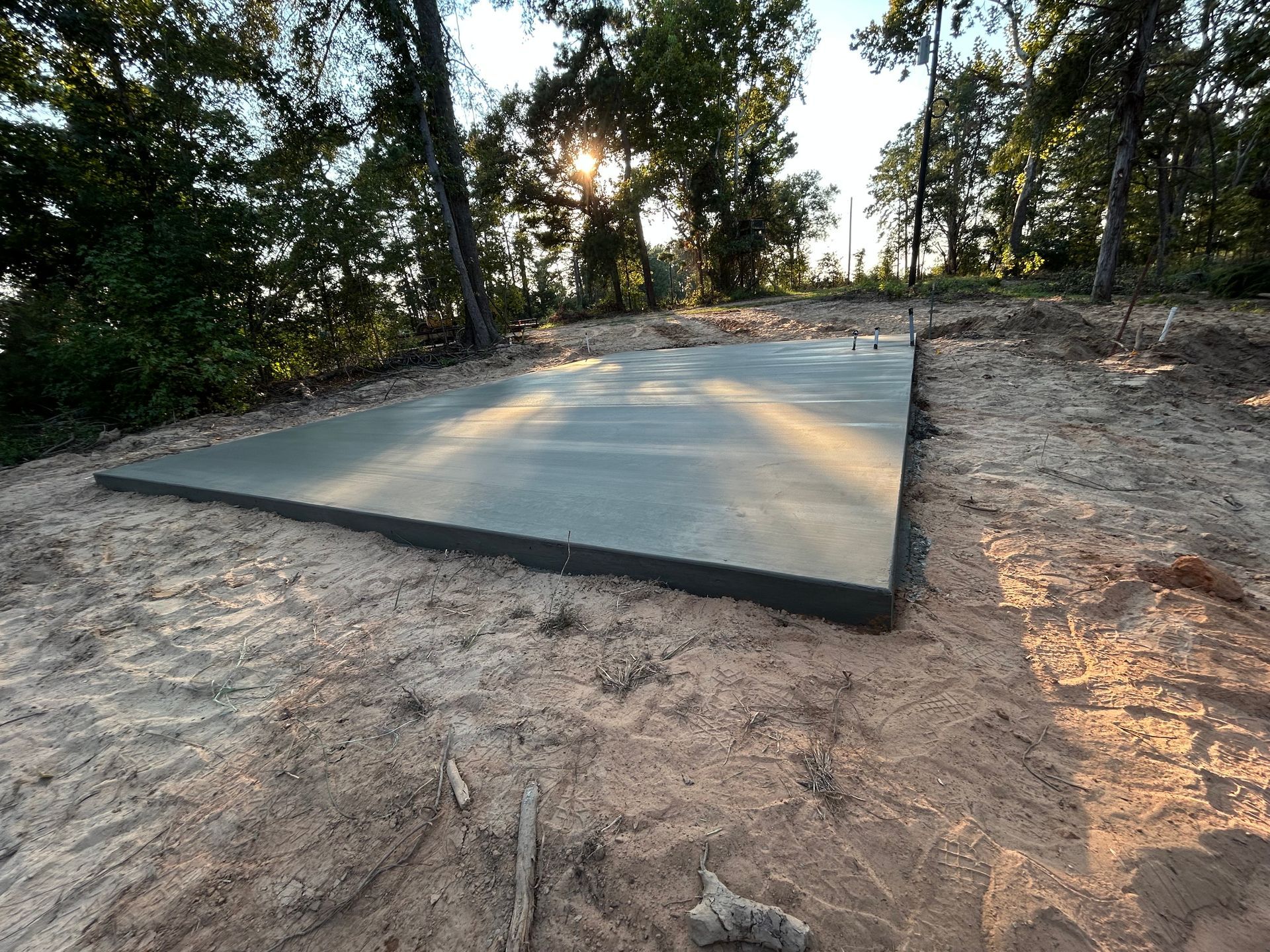 A freshly poured concrete foundation slab sits on a patch of cleared dirt in a wooded area during sunset.