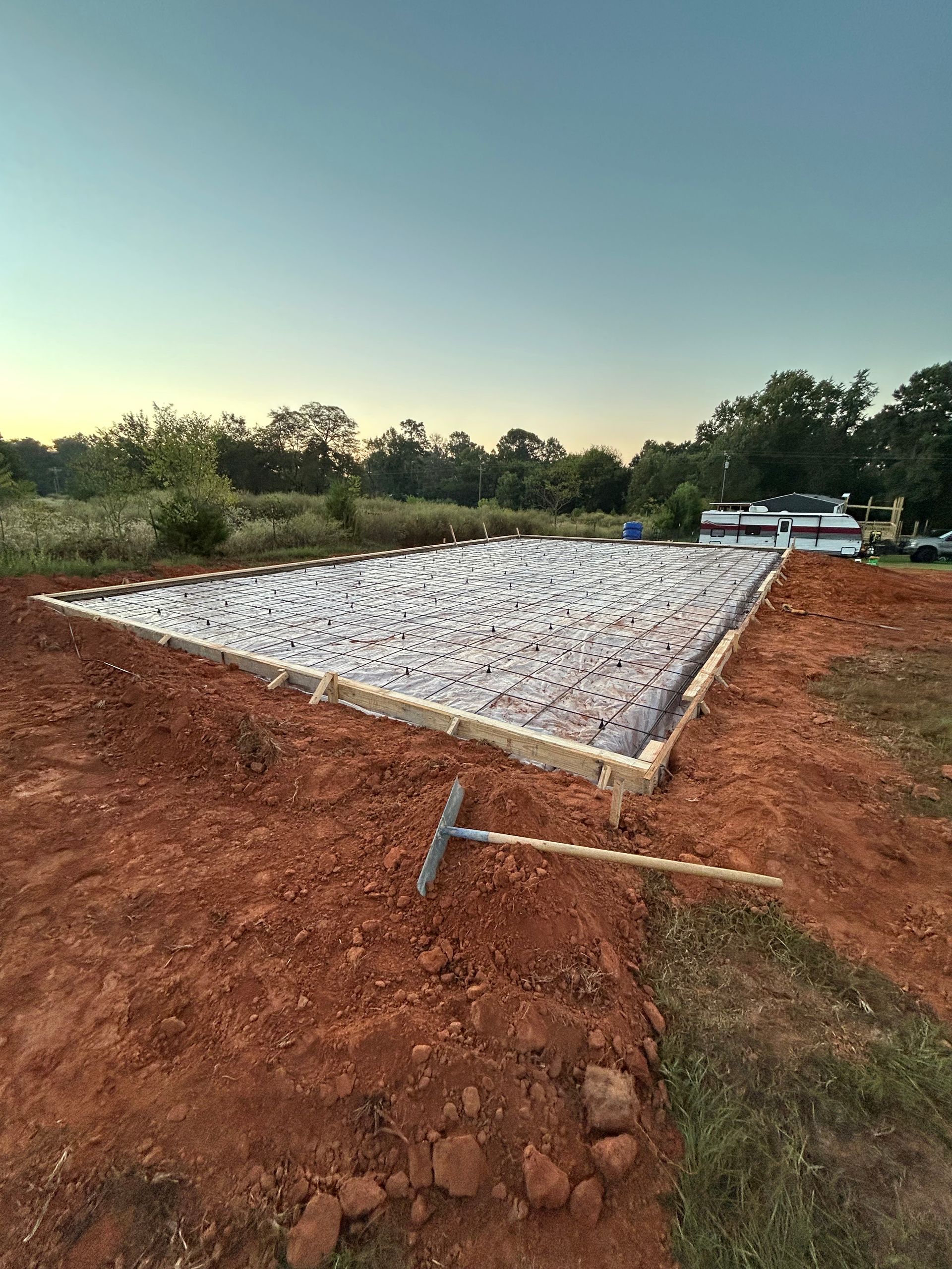 A gravel-filled rectangular construction foundation surrounded by red clay dirt at dusk, with a rake resting in the foreground.