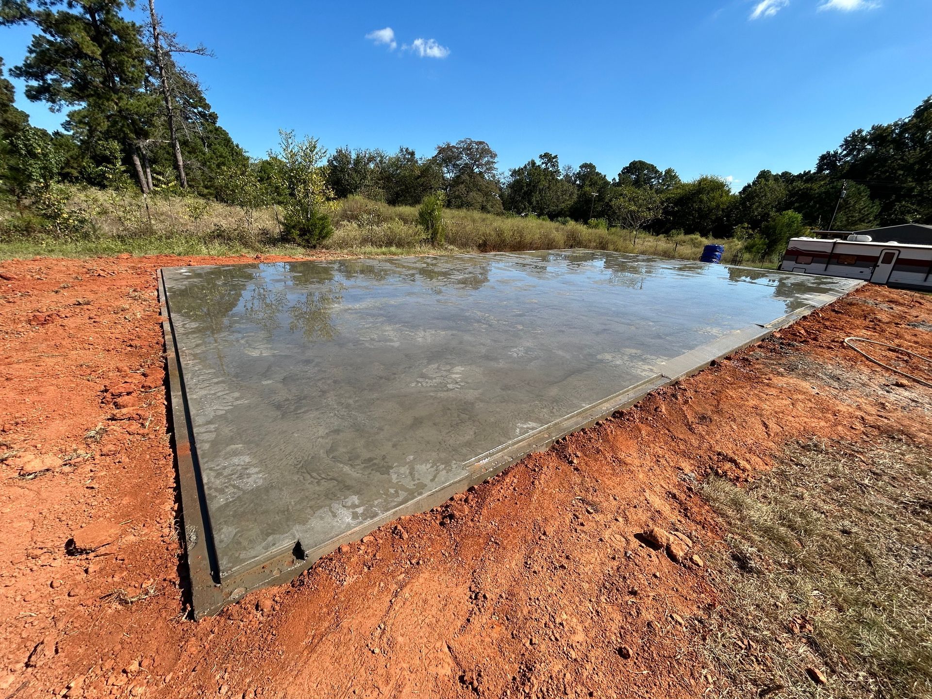 A freshly poured rectangular concrete slab surrounded by red dirt on a sunny day with trees in the background.