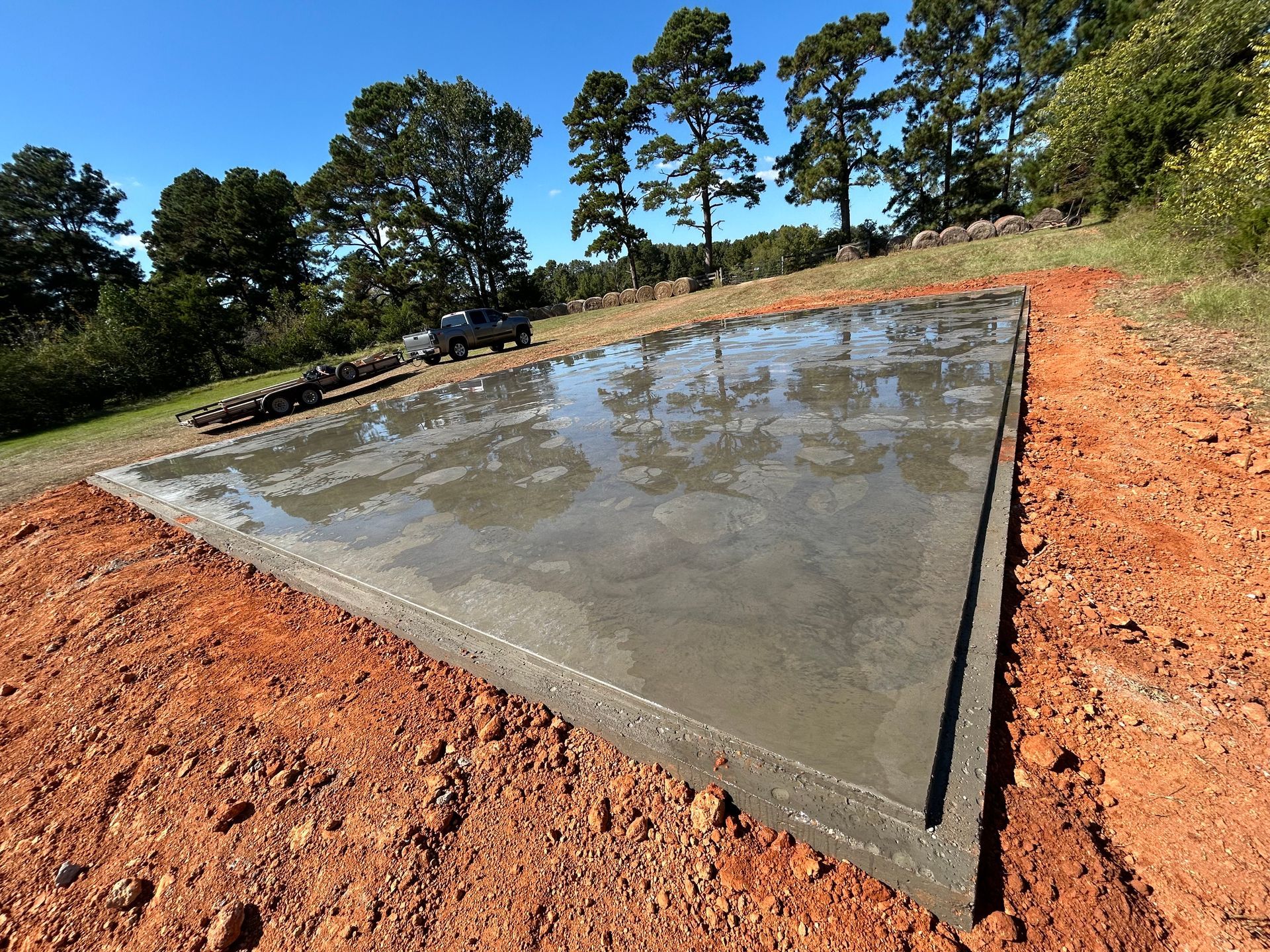 A freshly poured concrete slab foundation sits on red dirt, reflecting the sky and surrounding trees on a sunny day.