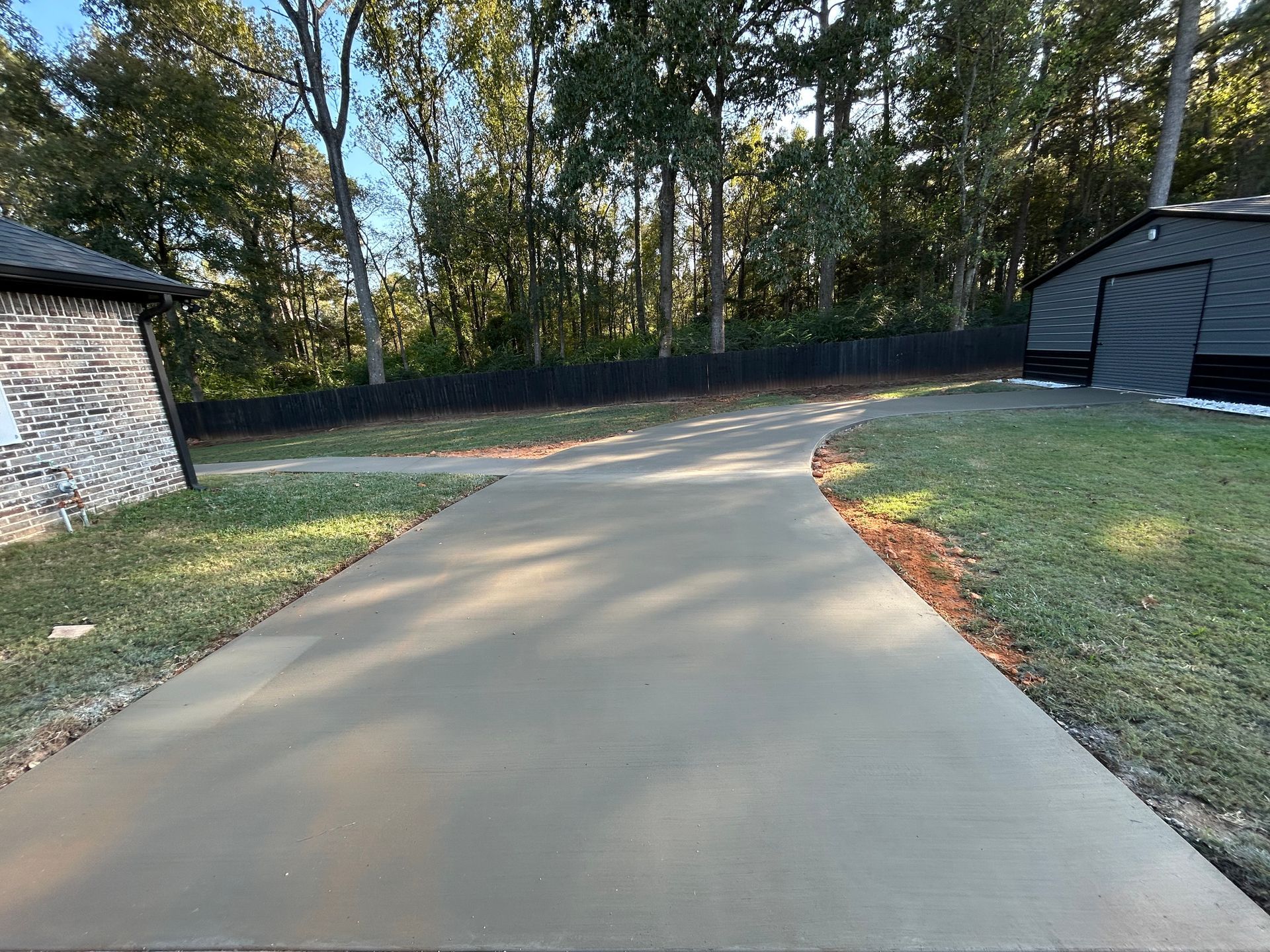 A smooth, newly poured concrete driveway leads between a stone-walled building and a dark metal shed toward a tree line.