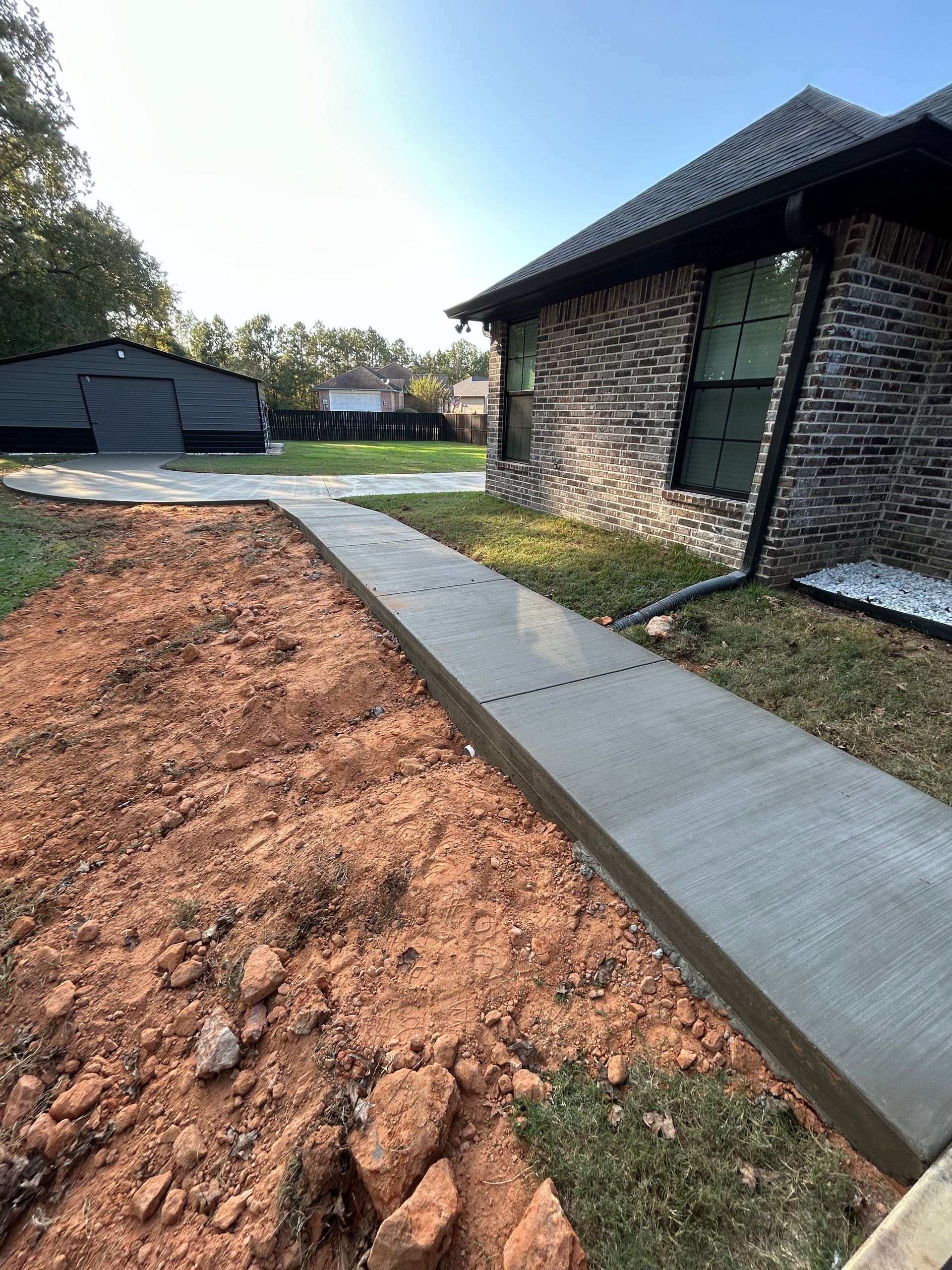 A newly poured concrete walkway runs alongside a brick house, leading toward a dark metal garage in the background.
