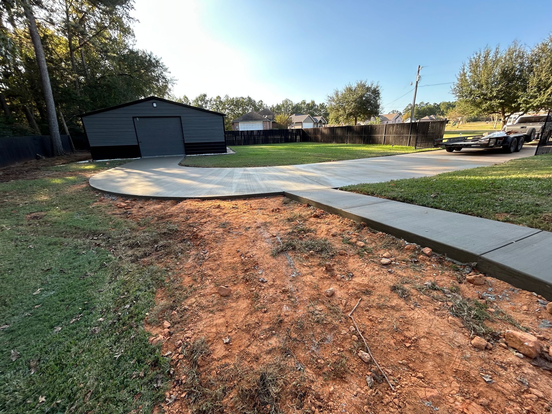 A newly poured concrete walkway and driveway leading to a dark gray garage in a grassy backyard with reddish soil.