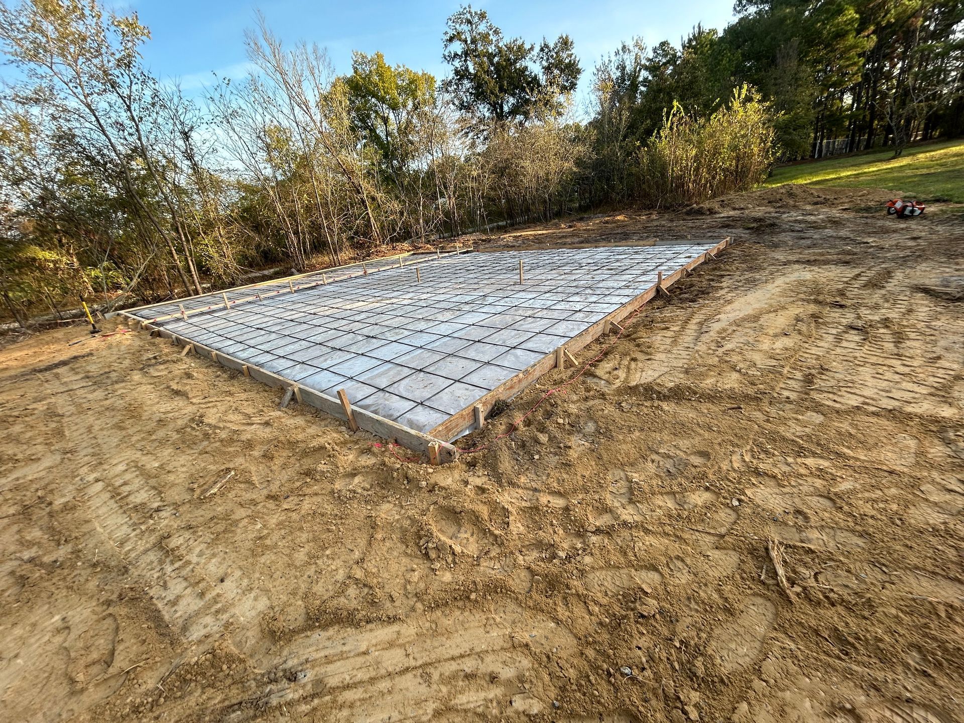 A rectangular concrete foundation reinforced with wire mesh sits in a clearing of dirt and grass.