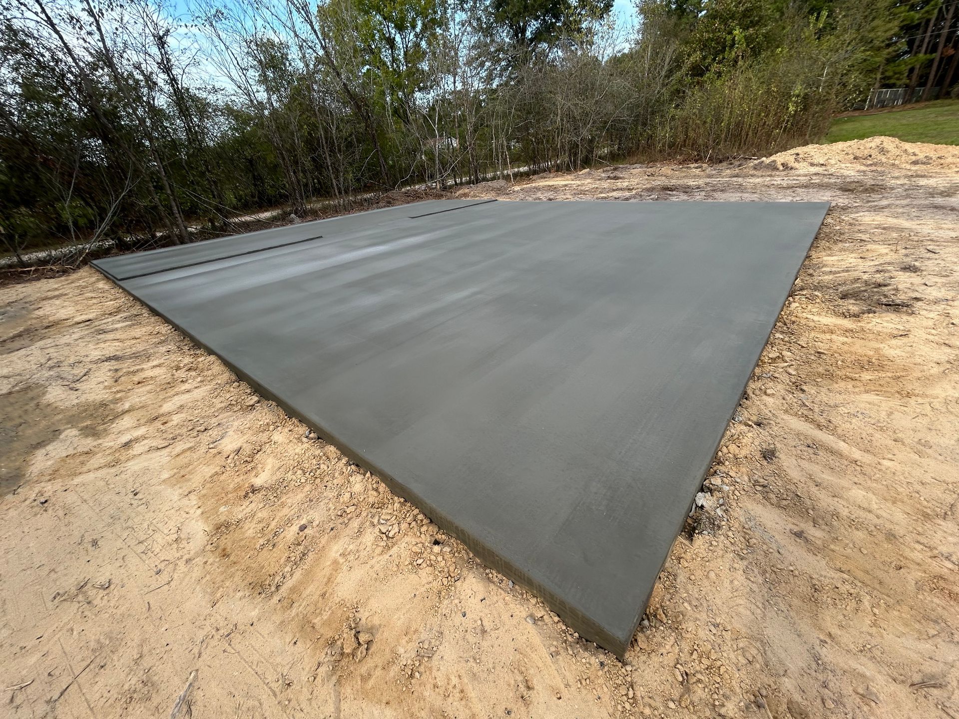 A newly poured, rectangular concrete slab sits on sandy ground surrounded by trees.