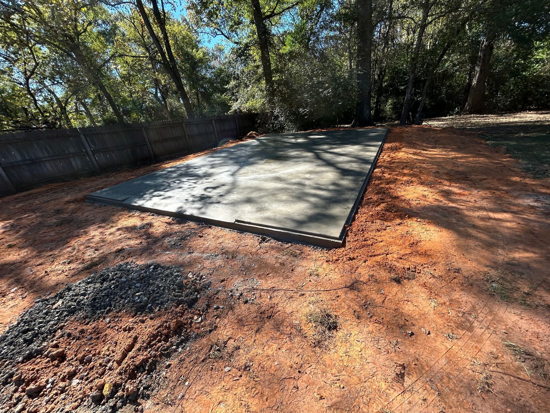 A newly poured concrete rectangular slab sits on red dirt in a wooded backyard near a wooden fence.