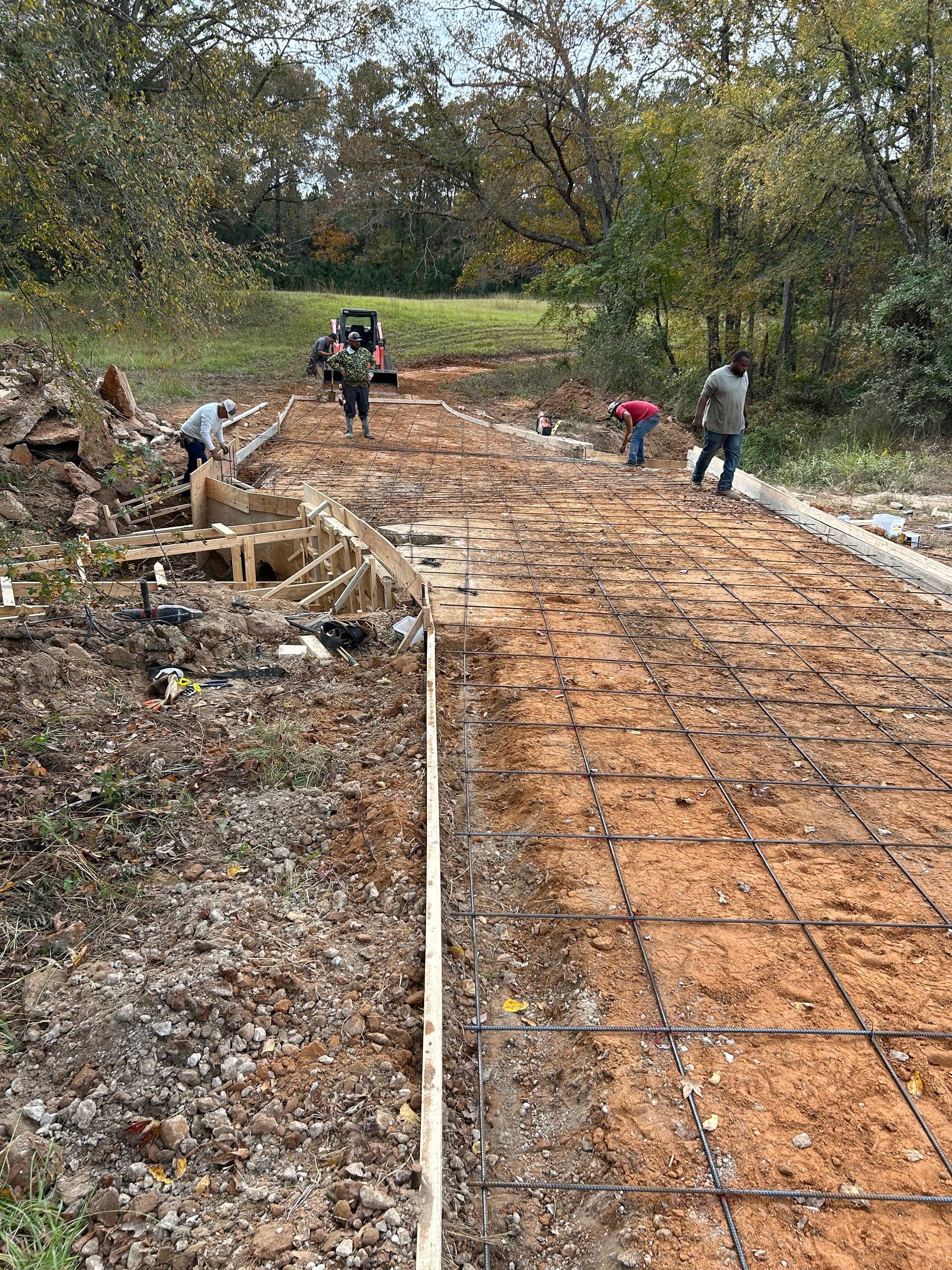 Workers prepare a dirt path lined with wooden forms and wire mesh for a concrete pour in a wooded area.