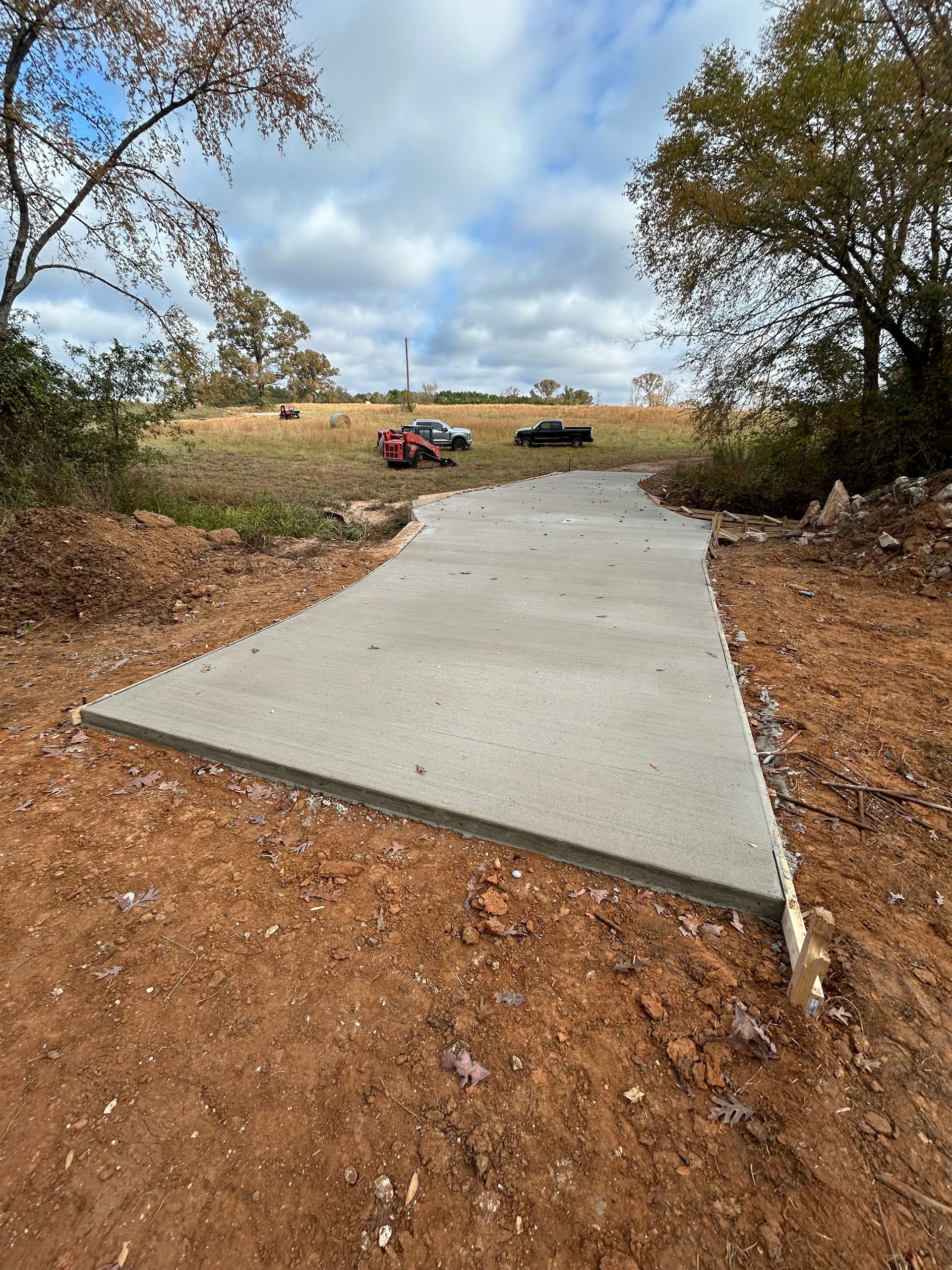 A newly poured concrete driveway curves through a rural, unlandscaped dirt lot under a cloudy blue sky.