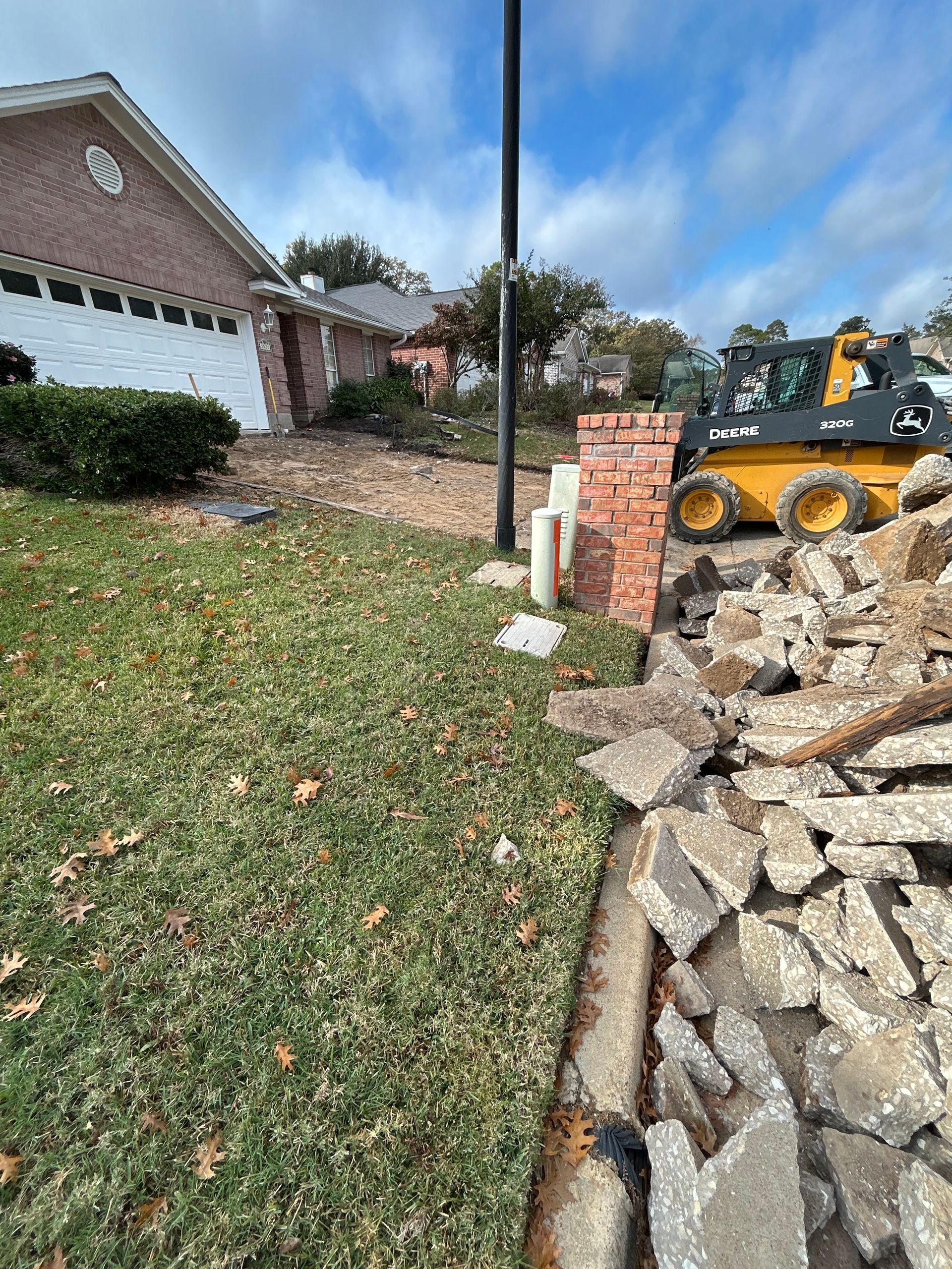 A yellow skid steer loader sits near a pile of broken concrete debris in the yard of a suburban brick house.