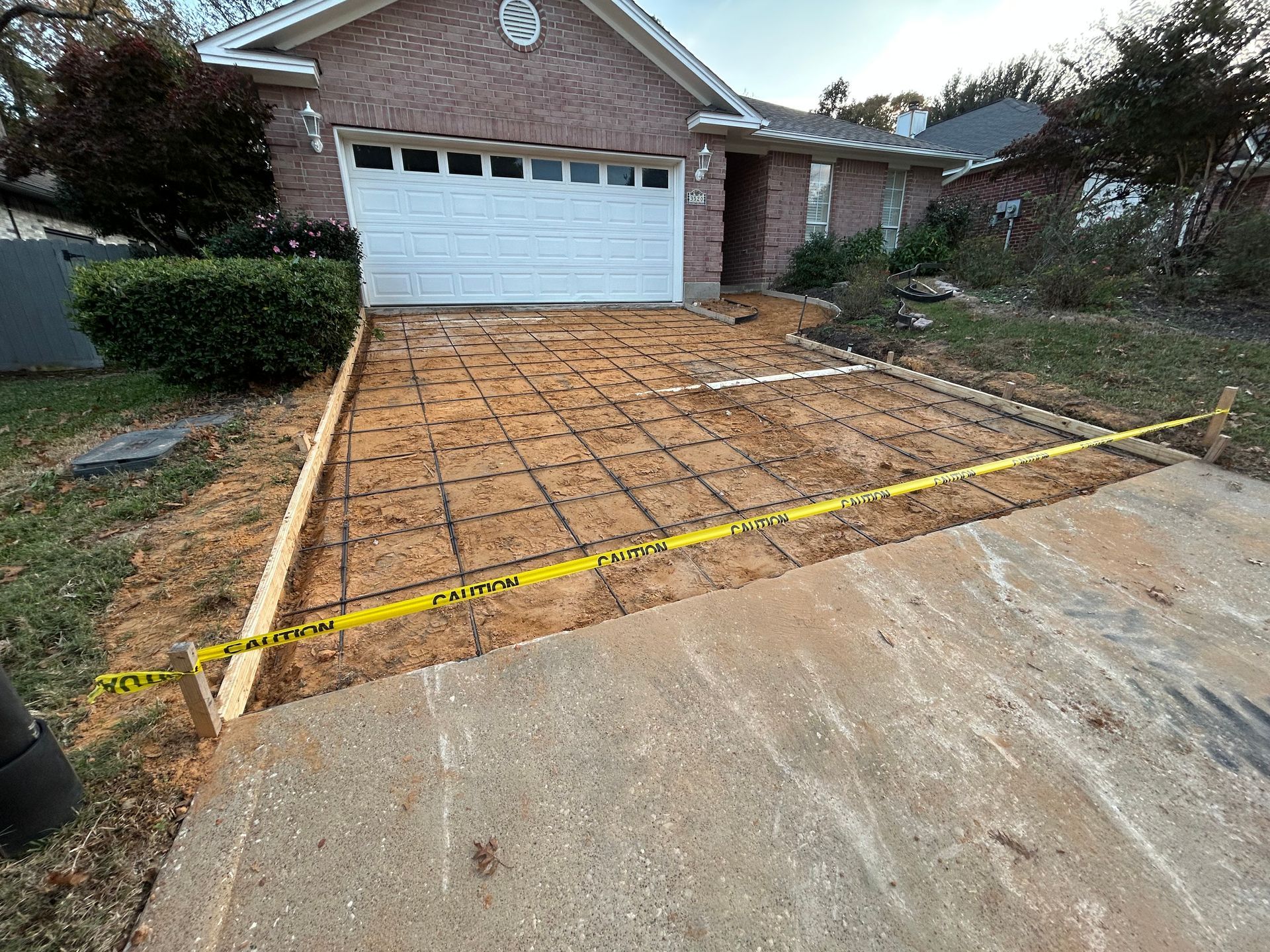A driveway under construction with wooden forms and metal grid reinforcement laid over dirt in front of a brick house.